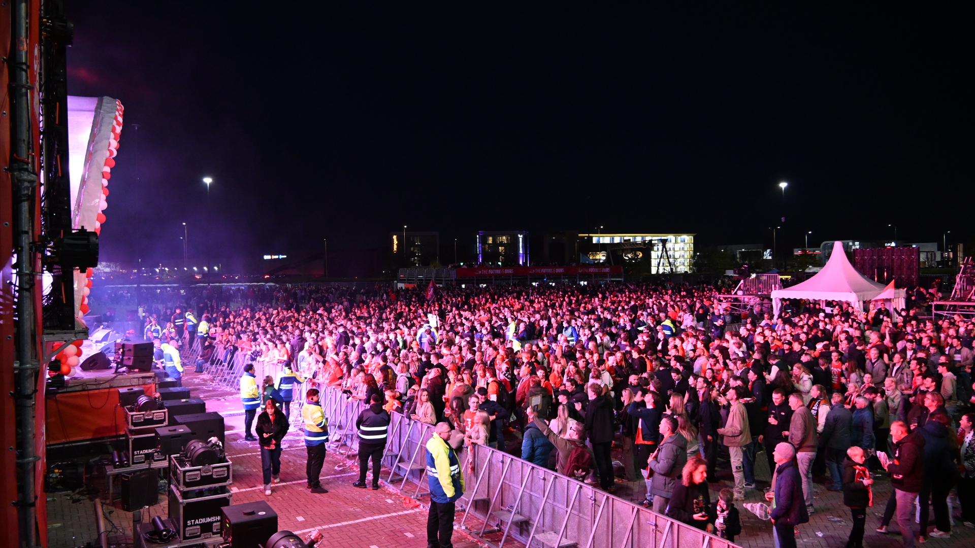 Groot publiek bij een avondconcert, met beveiligers voor de hekken en een verlichte tent op de achtergrond.