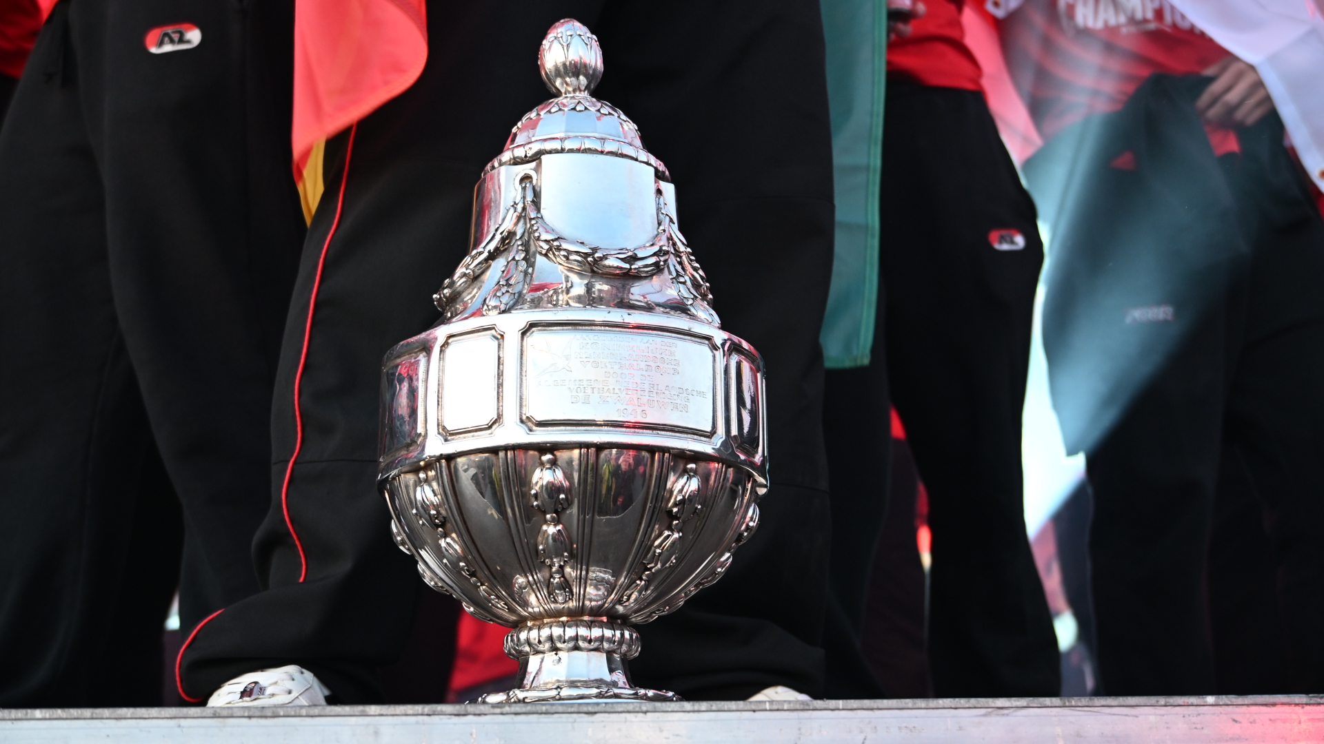 Een zilveren sporttrofee met gravure, omringd door mensen in sportkleding met het logo van AZ.