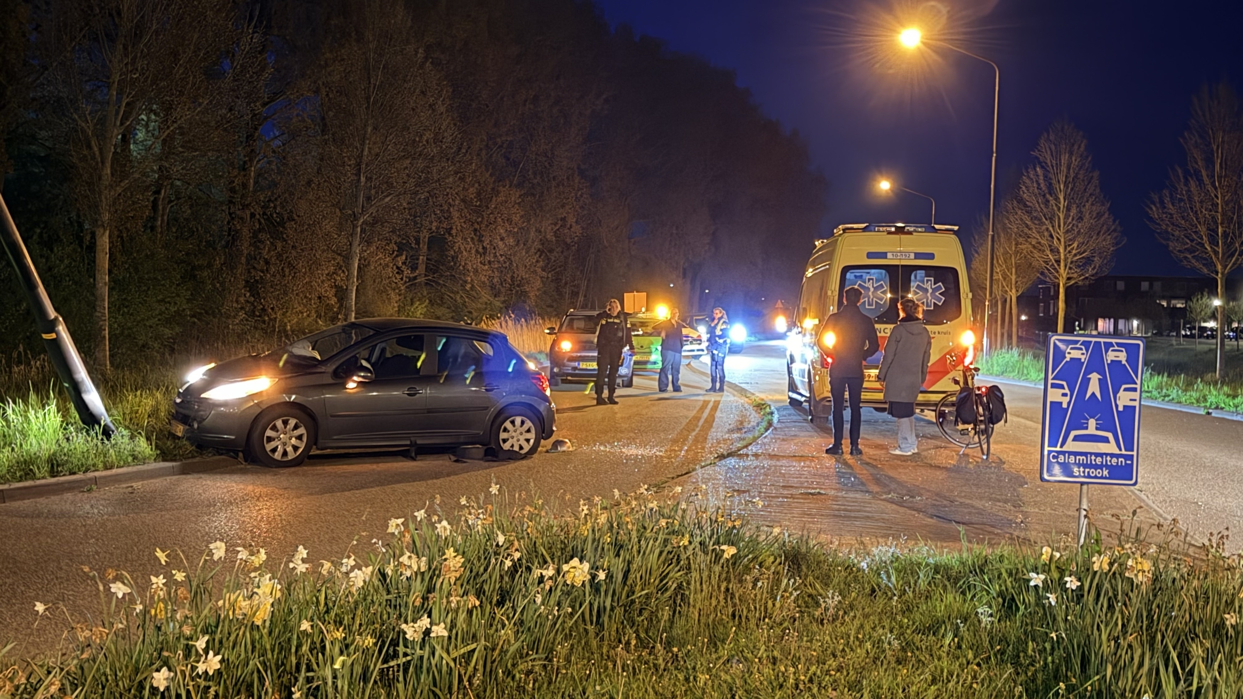 Nachtelijke verkeersscene met een auto die tegen een paal botst, omgeven door hulpverleners en een ambulance, naast een calamiteitenstrookbord.