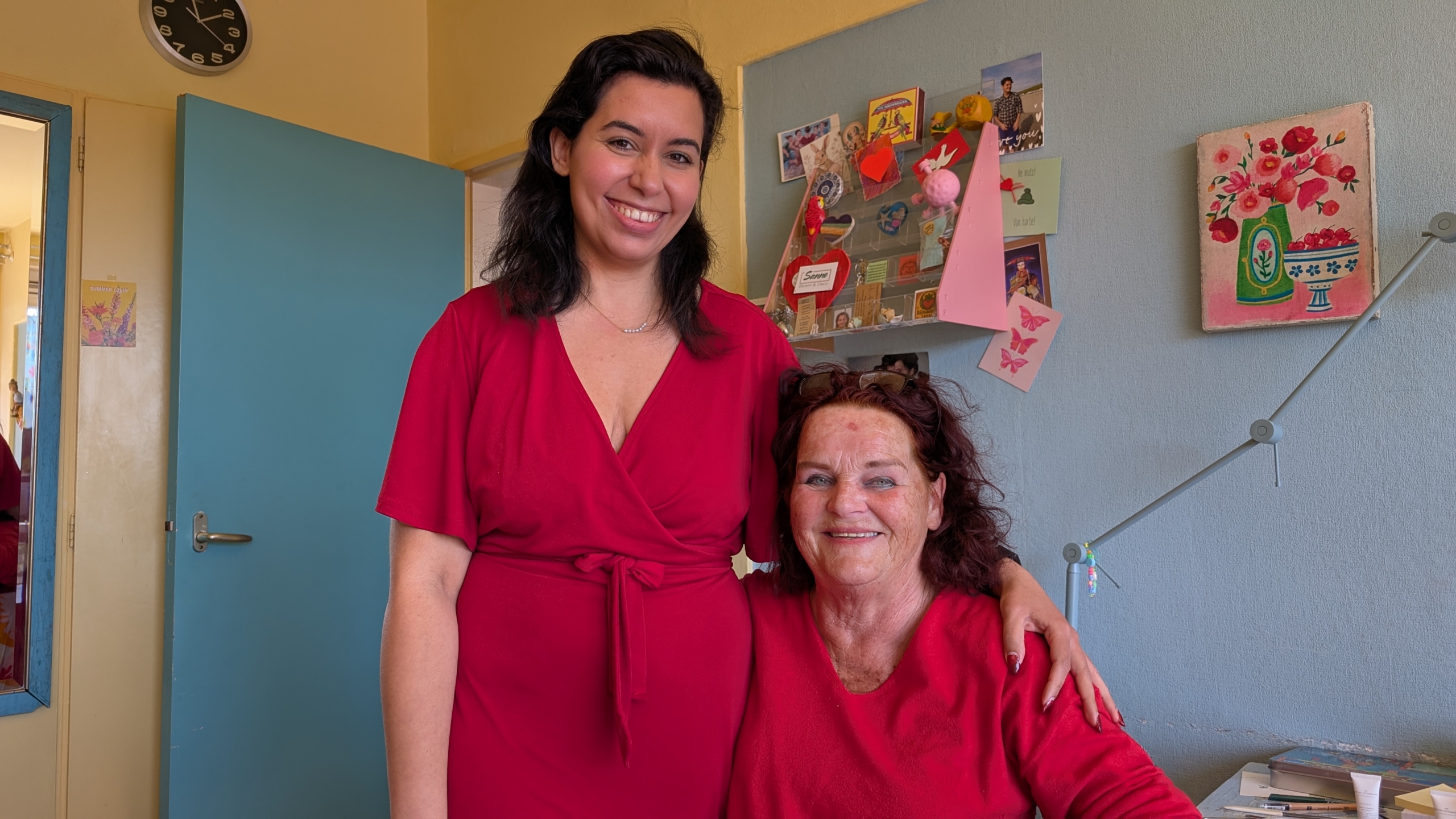Twee vrouwen glimlachen, beiden in een rode outfit, in een kamer met kleurrijke decoraties aan de muur.