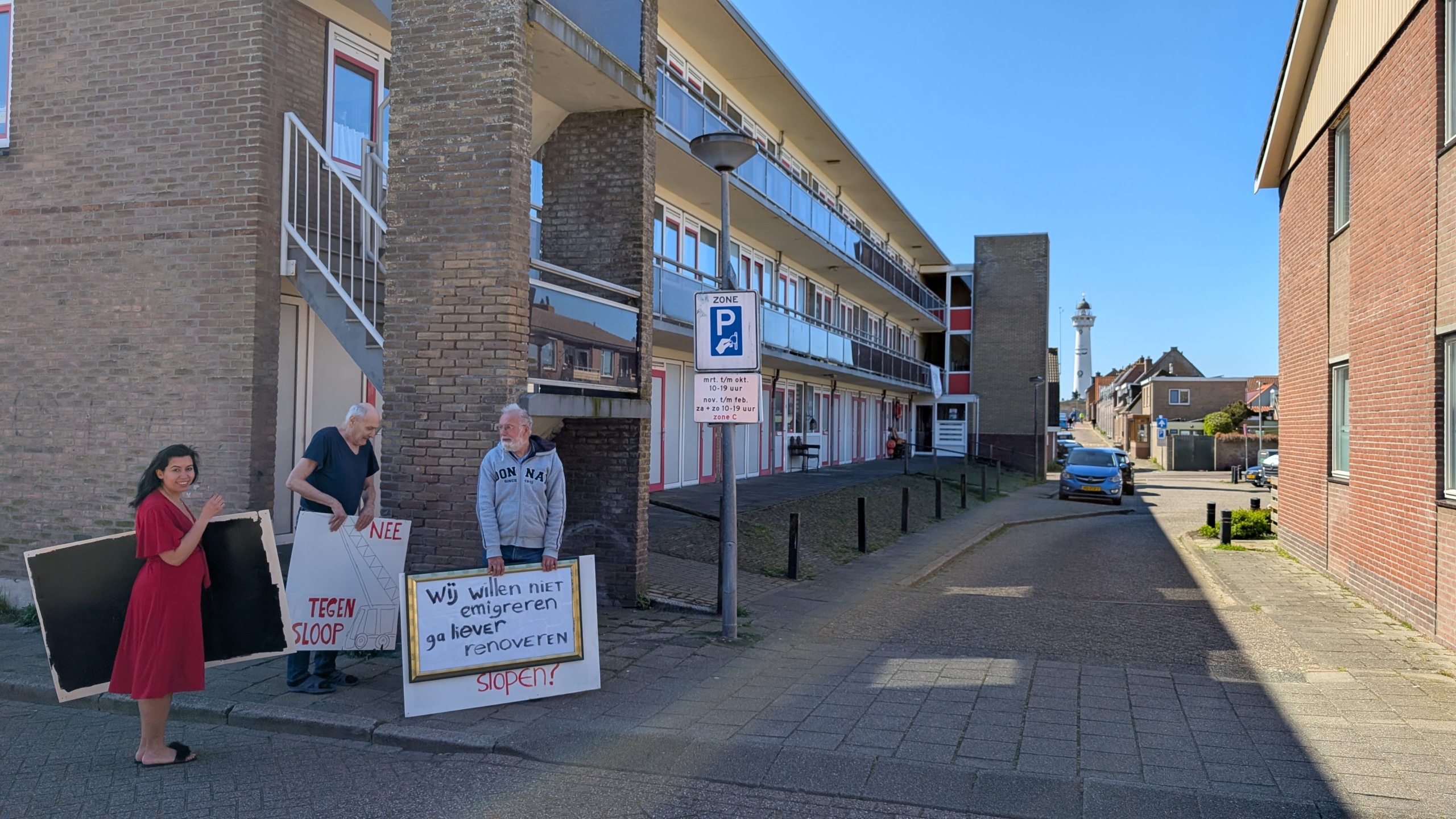 Mensen protesteren met borden tegen sloop voor een appartementsgebouw, met een straat en een vuurtoren op de achtergrond.
