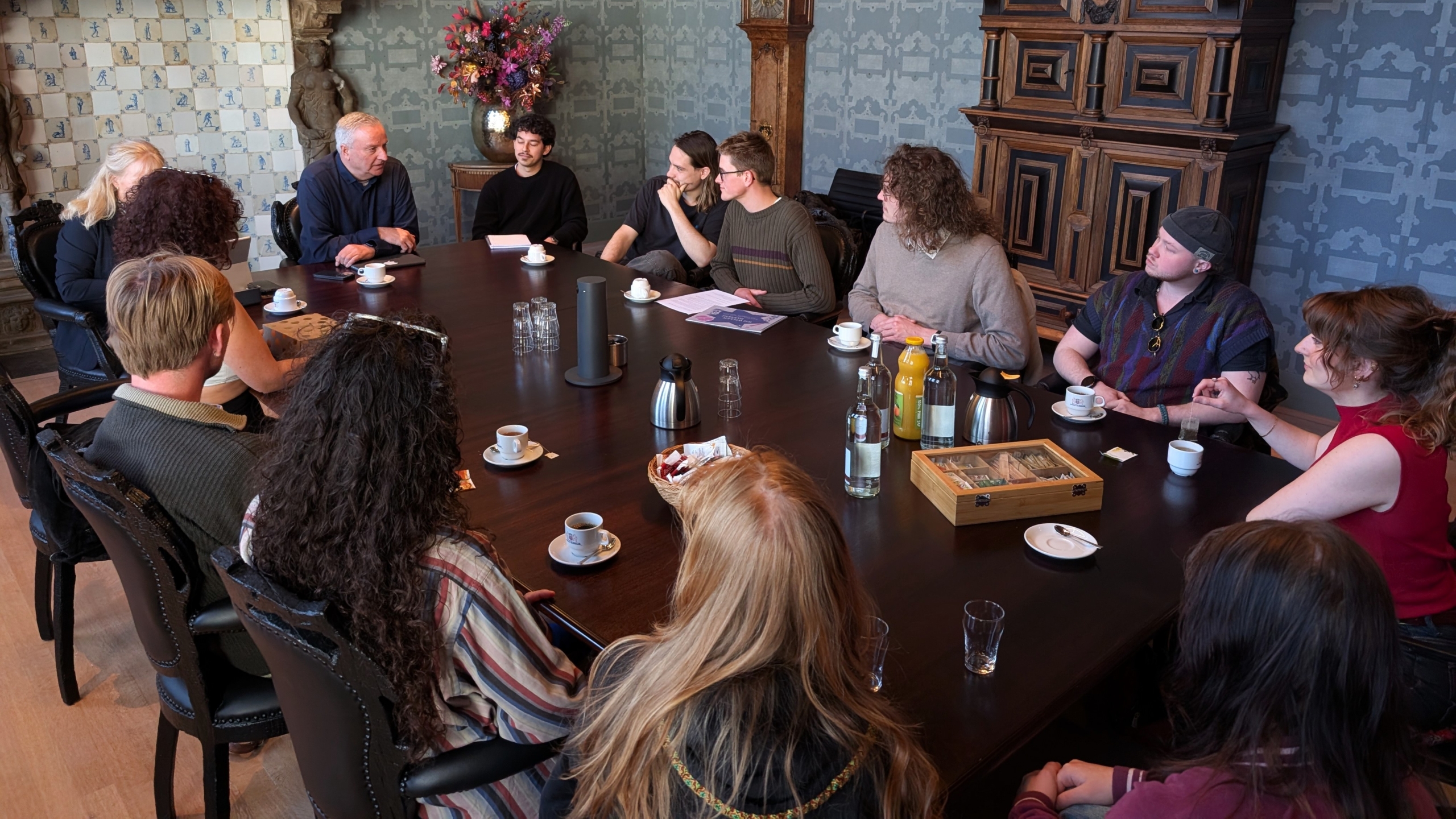 Mensen zitten rond een grote tafel in een vergaderruimte met koffie en drankjes, terwijl een oudere man spreekt.