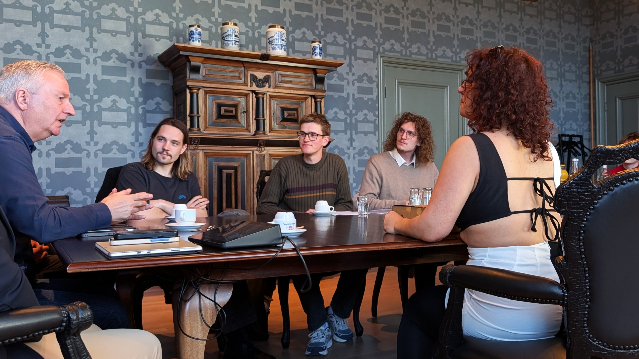 Vijf mensen in gesprek rond een tafel in een kamer met een antiek dressoir en traditionele decoratie.