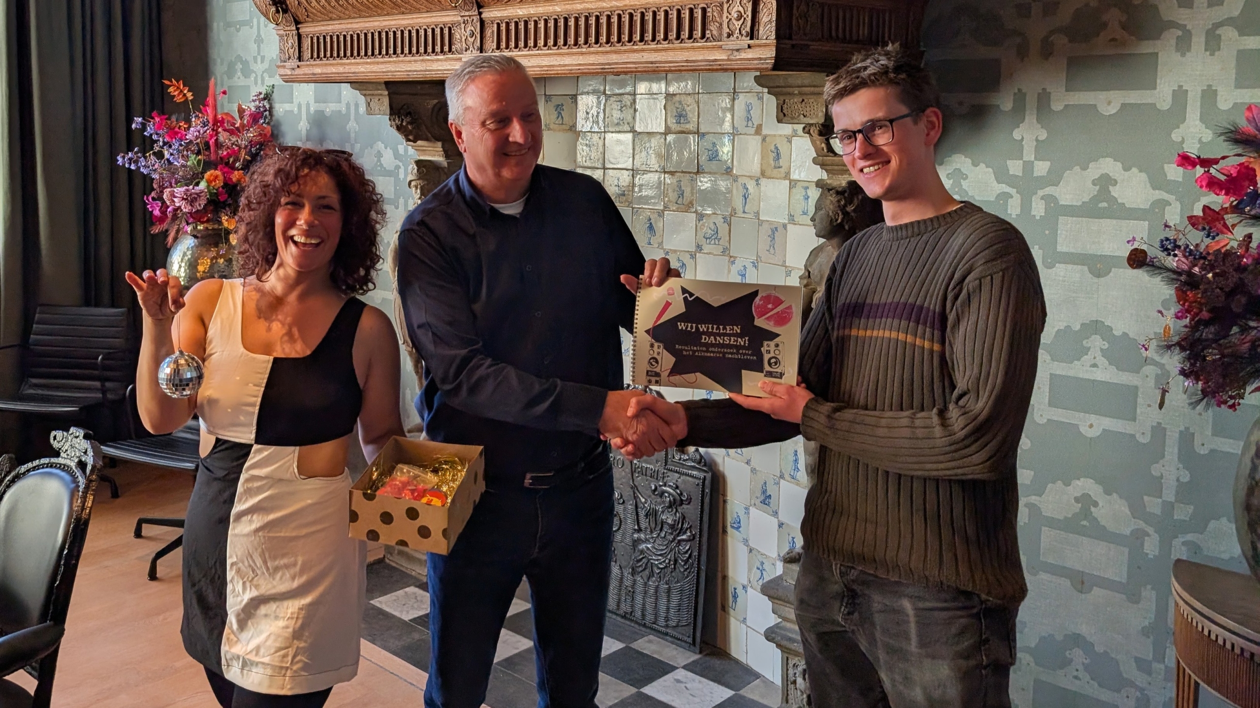 Drie mensen poseren lachend; een man schudt handen met een jongere man terwijl een vrouw een doos en een kerstbal vasthoudt.