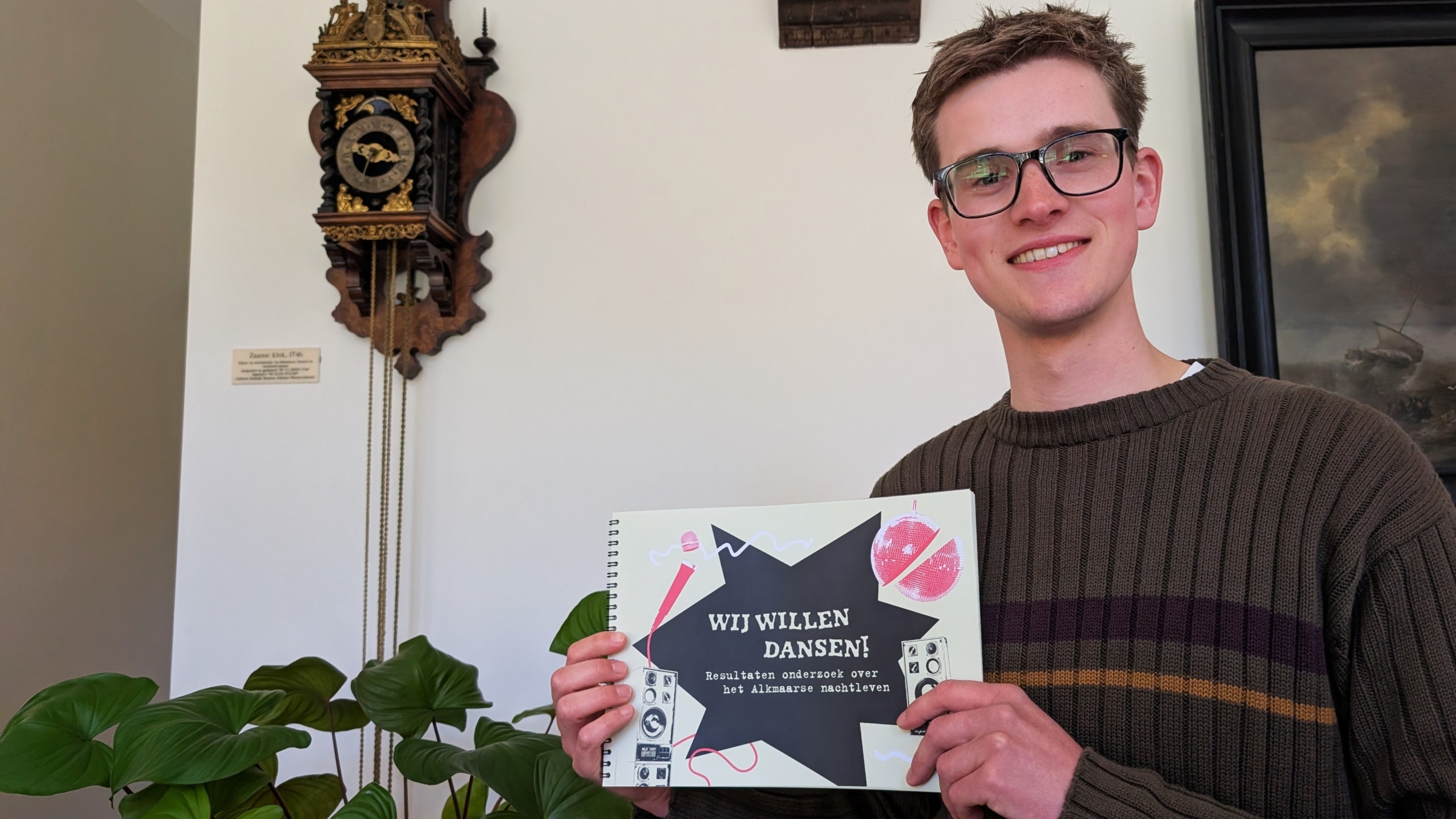 Man met bril houdt een boek vast met de tekst "WIJ WILLEN DANSEN!" in een kamer met een klok en schilderij.