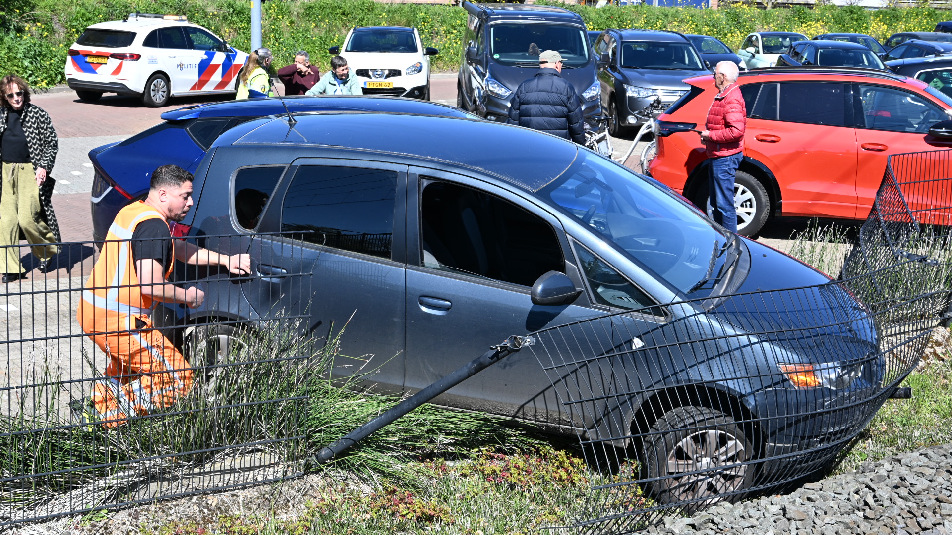 Auto tegen schuin hek met man in oranje vest die de deur opent. Politiewagen en omstanders op de achtergrond.