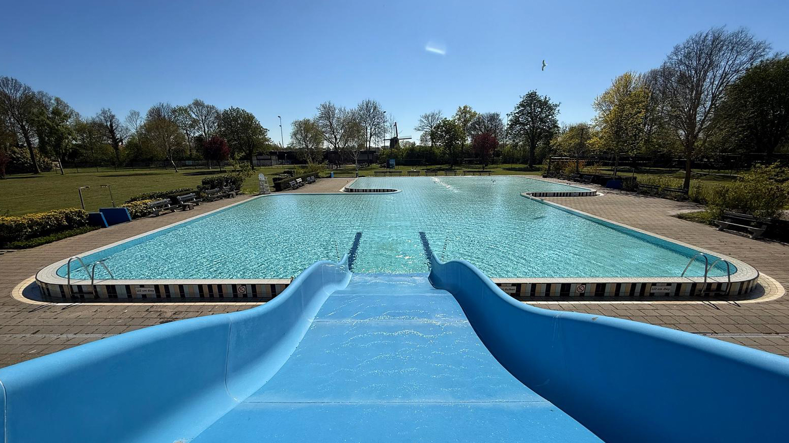 Een leeg buitenzwembad met een blauwe glijbaan in een parkachtige omgeving op een zonnige dag.