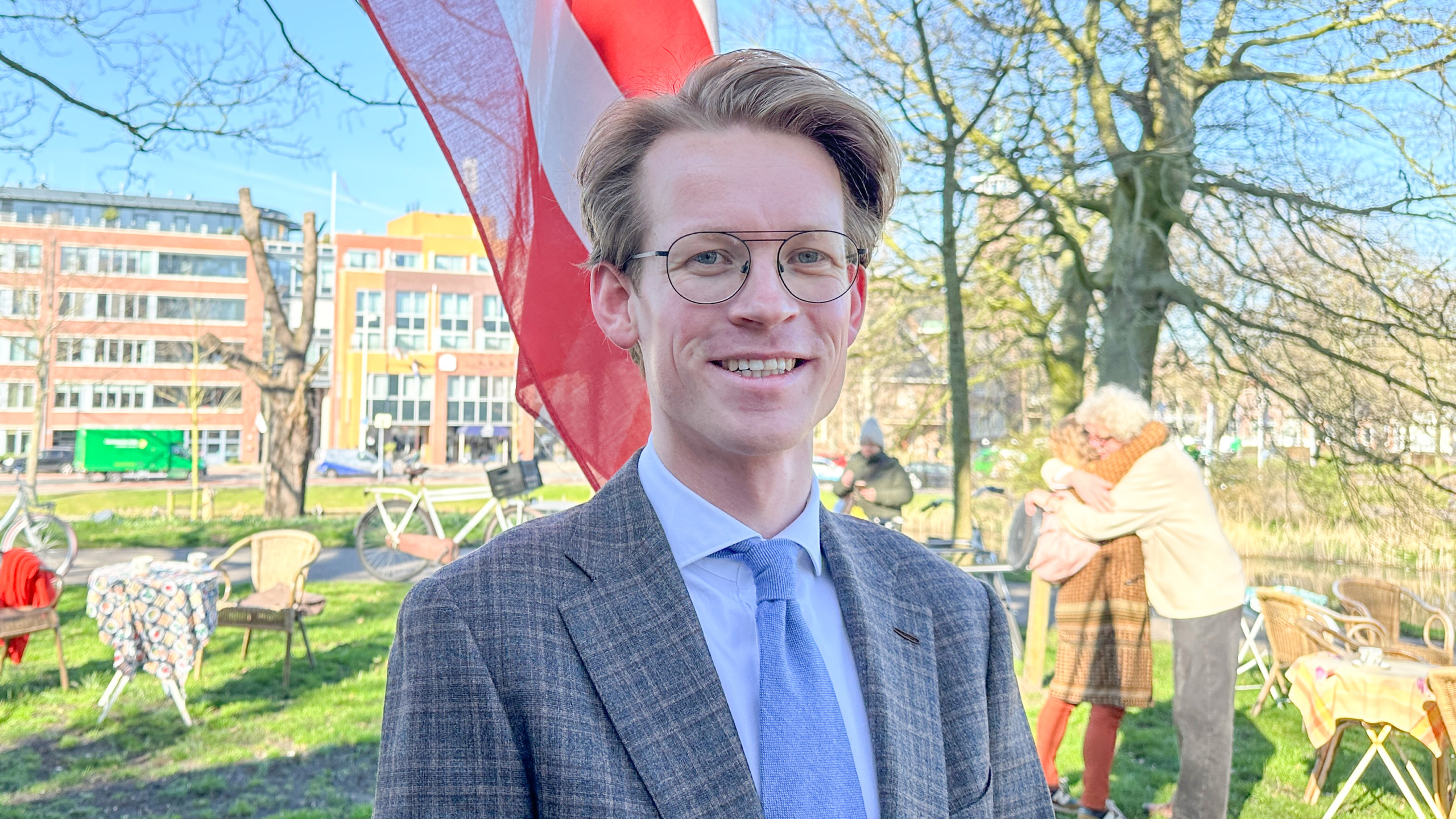 Man in een pak met een blauwe stropdas en een bril, staand buiten bij tafels en stoelen, met een vlag op de achtergrond.