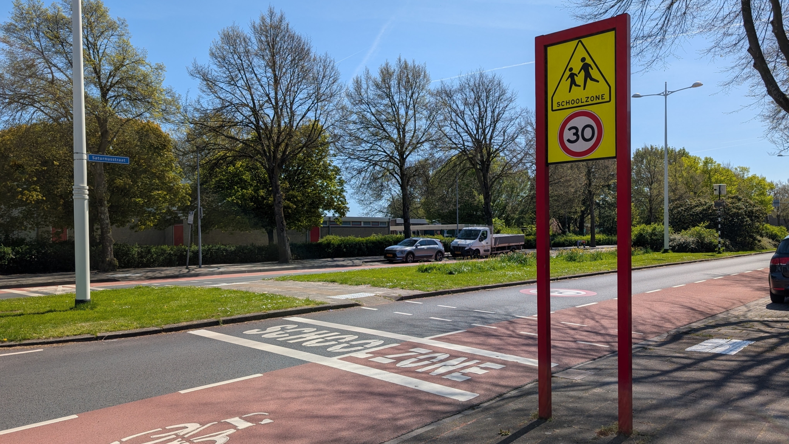 Verkeersbord met "Schoolzone" en maximumsnelheid van 30 km/u, langs een rustige straat met bomen en geparkeerde auto's.