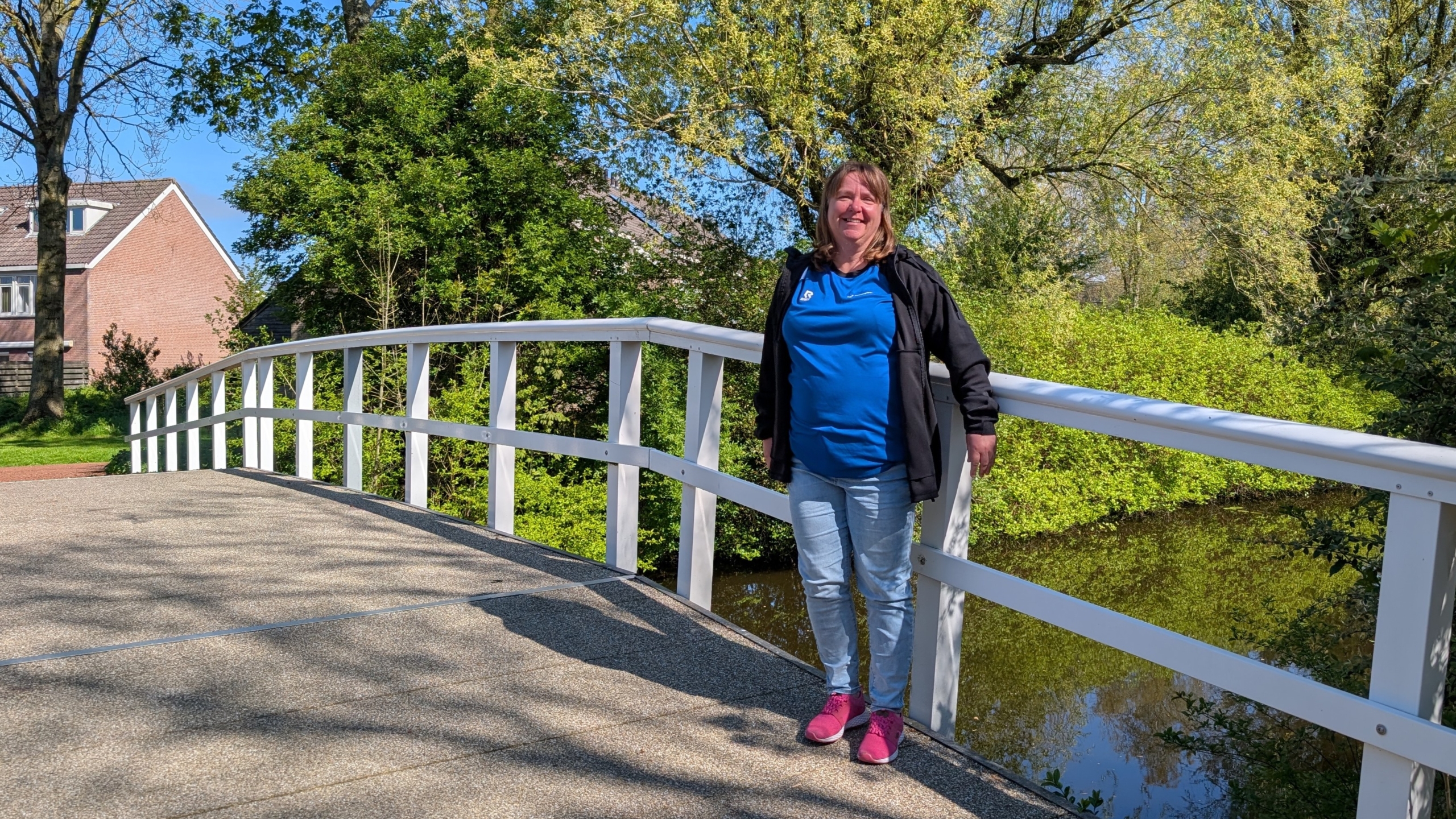 Vrouw in blauwe blouse en roze schoenen leunt op een witte brugleuning, met groene bomen en huizen op de achtergrond.