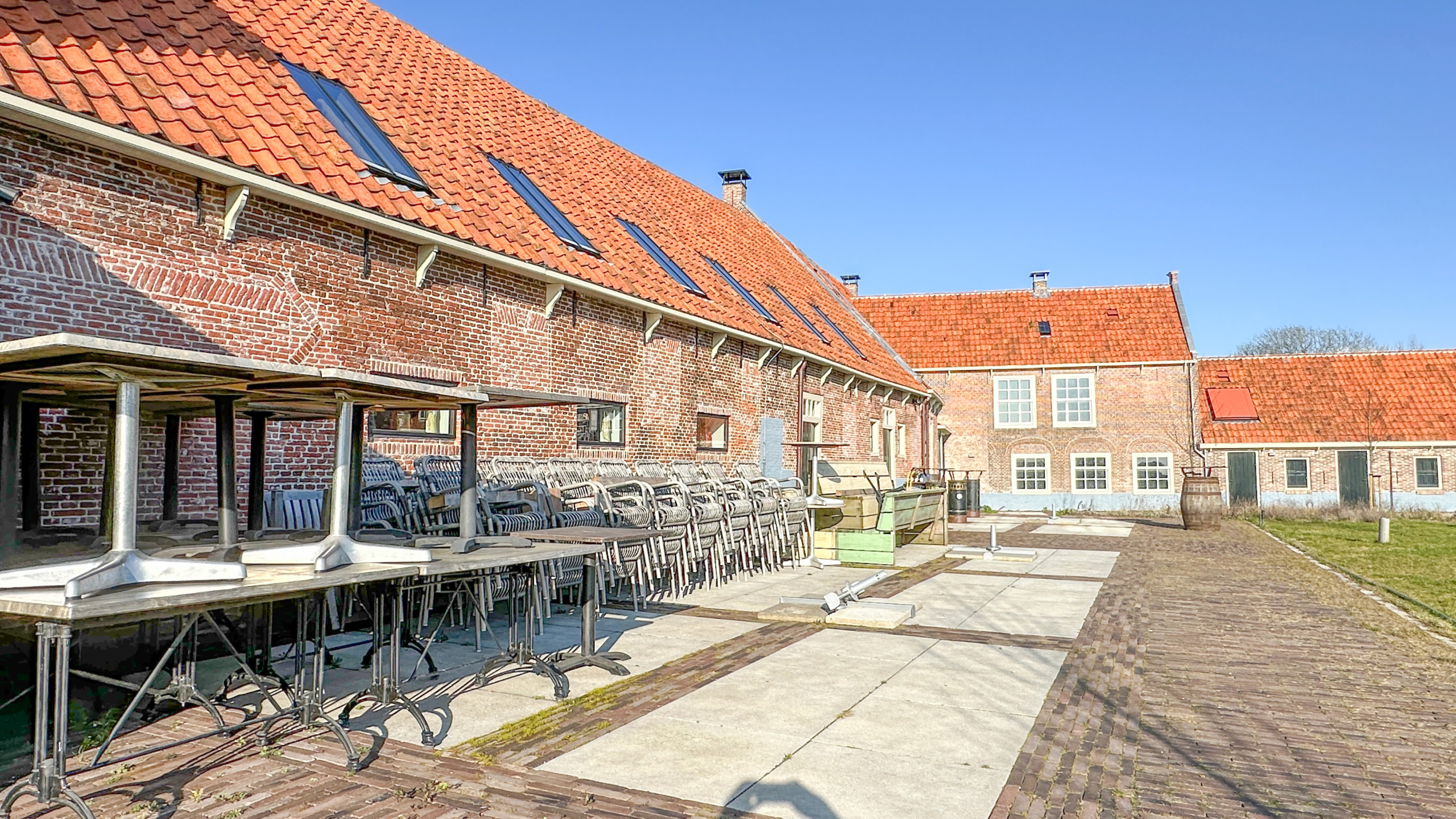 Bakstenen gebouw met rode dakpannen, omgeven door gestapelde terrasstoelen en omgekeerde tafels op een betegelde binnenplaats.
