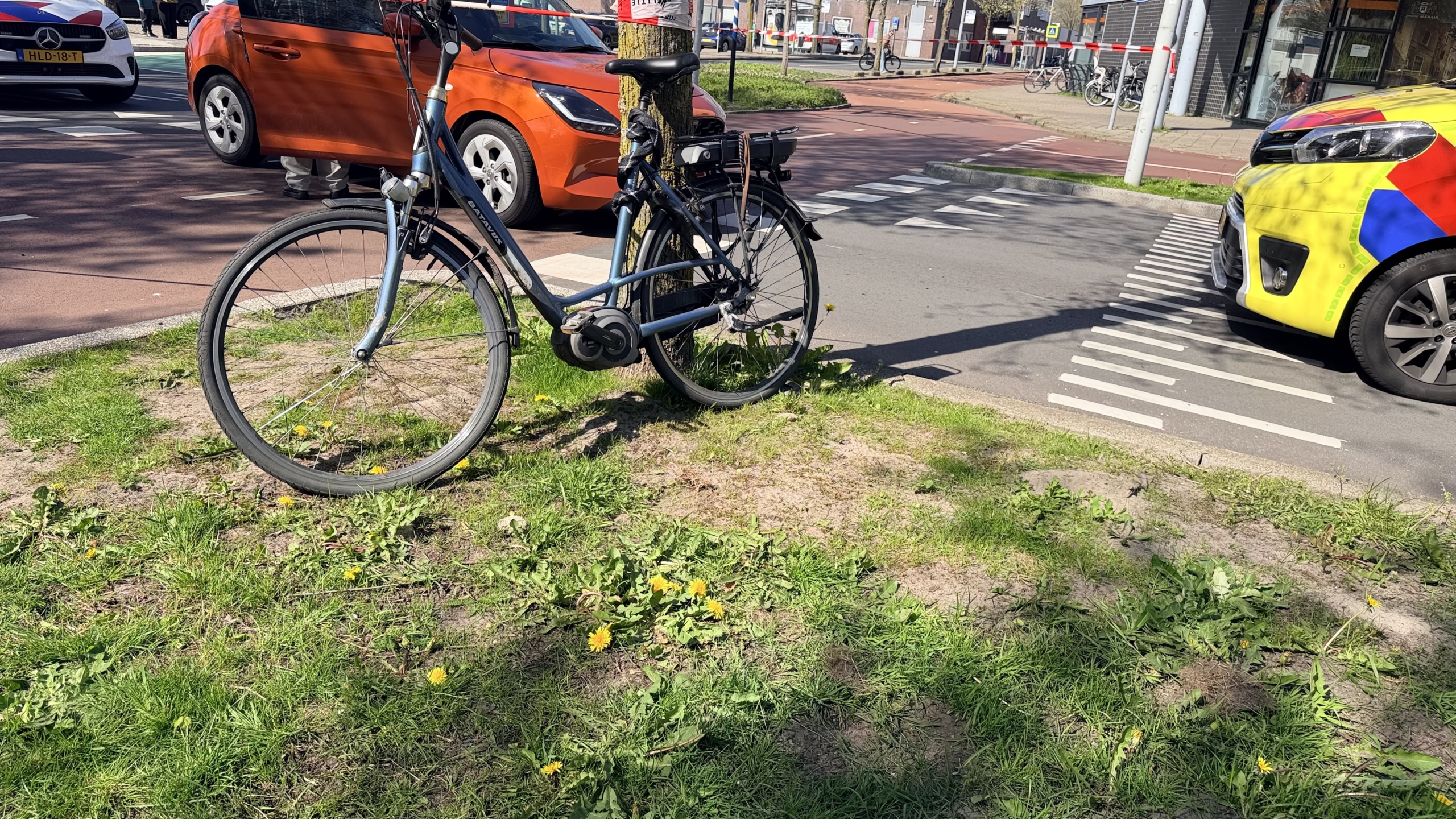 Een fiets is vastgebonden aan een boom naast een weg, naast een felgele auto en een oranje auto.