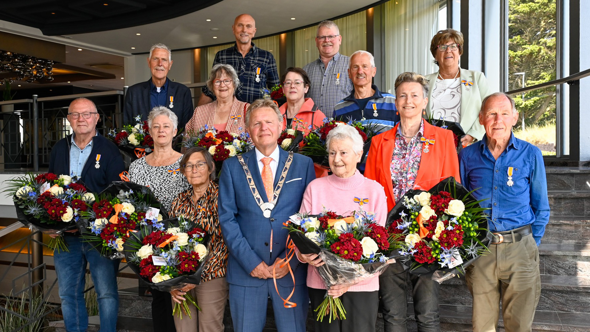Een groep mensen die lintjes en grote boeketten bloemen vasthouden, staat samen met een persoon in een ambtsketen op een trap binnenshuis.