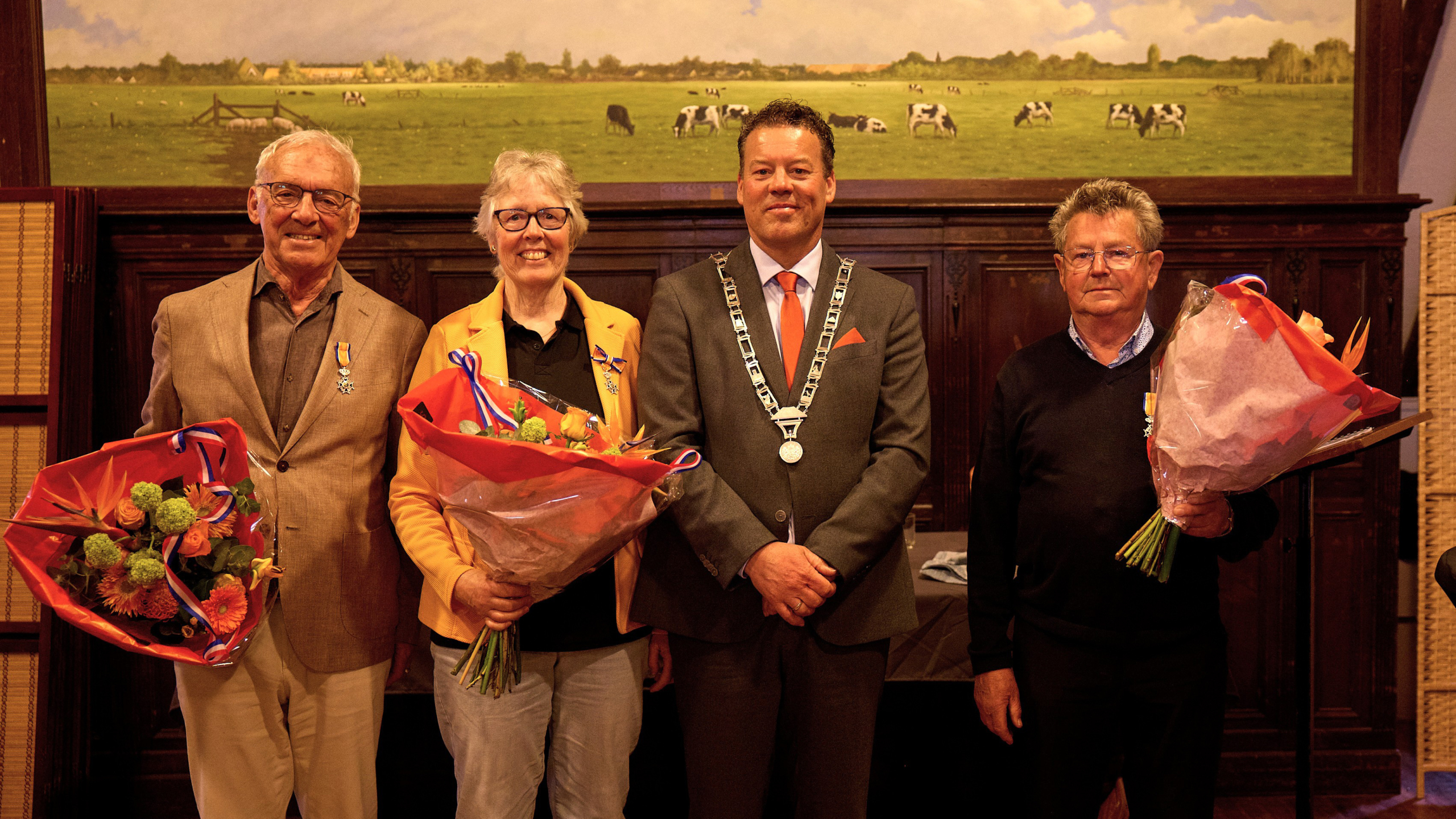 Vier mensen staan naast elkaar; drie dragen lintjes en houden boeketten vast, terwijl de vierde een keten om de hals draagt. Op de achtergrond is een schilderij van een groene weide met koeien te zien.