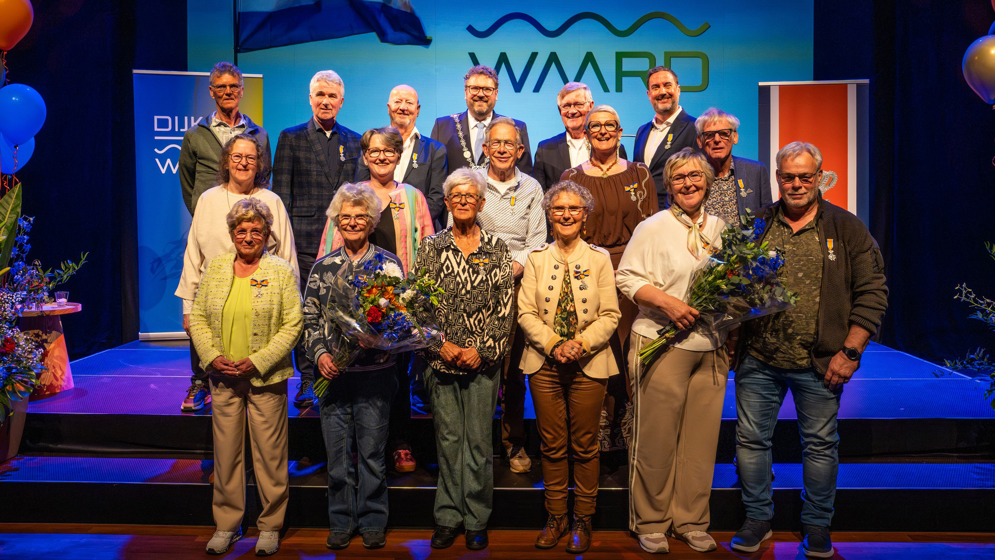 Groep mensen poseert op een podium, sommigen dragen onderscheidingen; bloemen en banners zijn zichtbaar op de achtergrond.