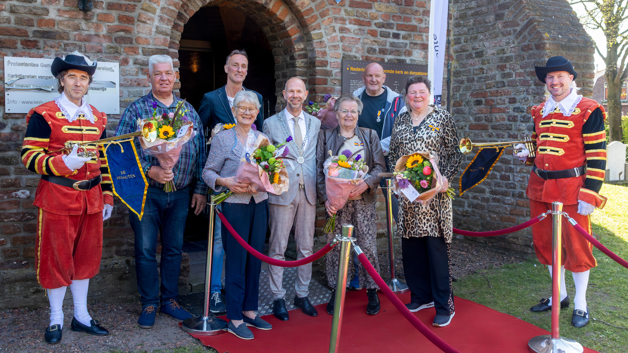 Een groep mensen poseert met bloemen voor een stenen gebouw, met twee trompettisten in rood uniform.