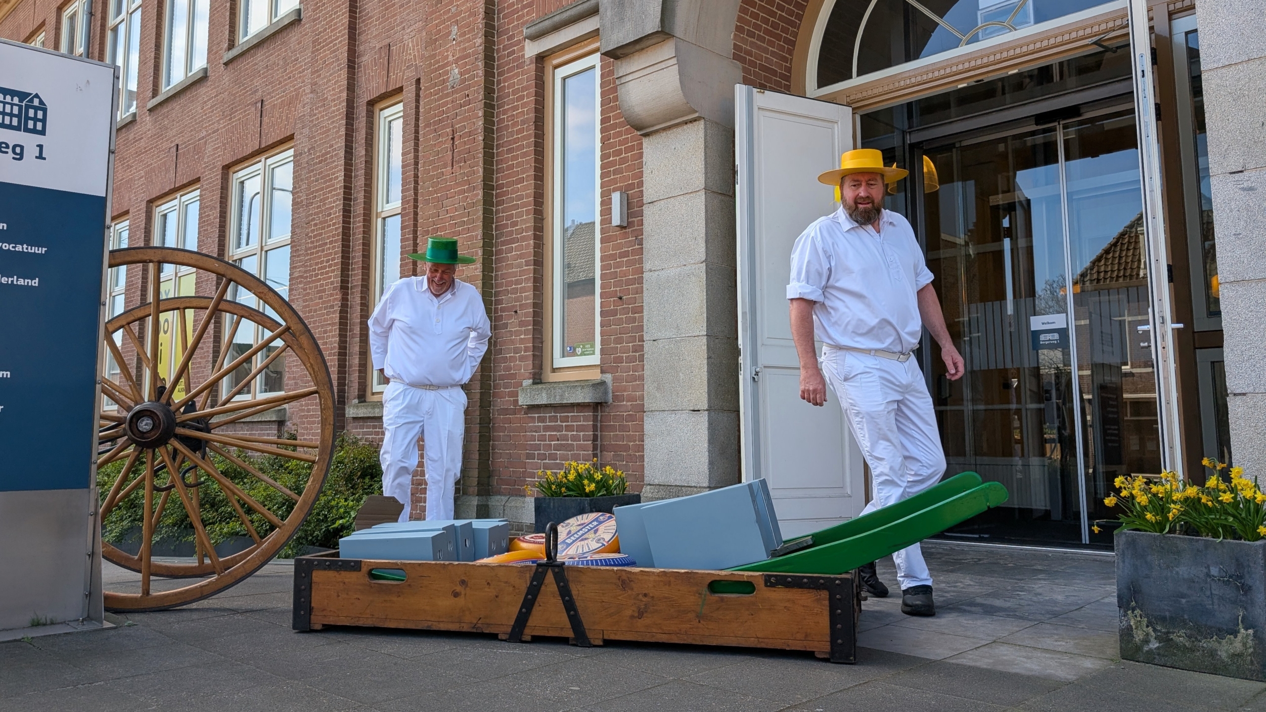 Twee mannen in traditionele kleding met gekleurde hoeden staan voor een gebouw, naast een kar met kazen.