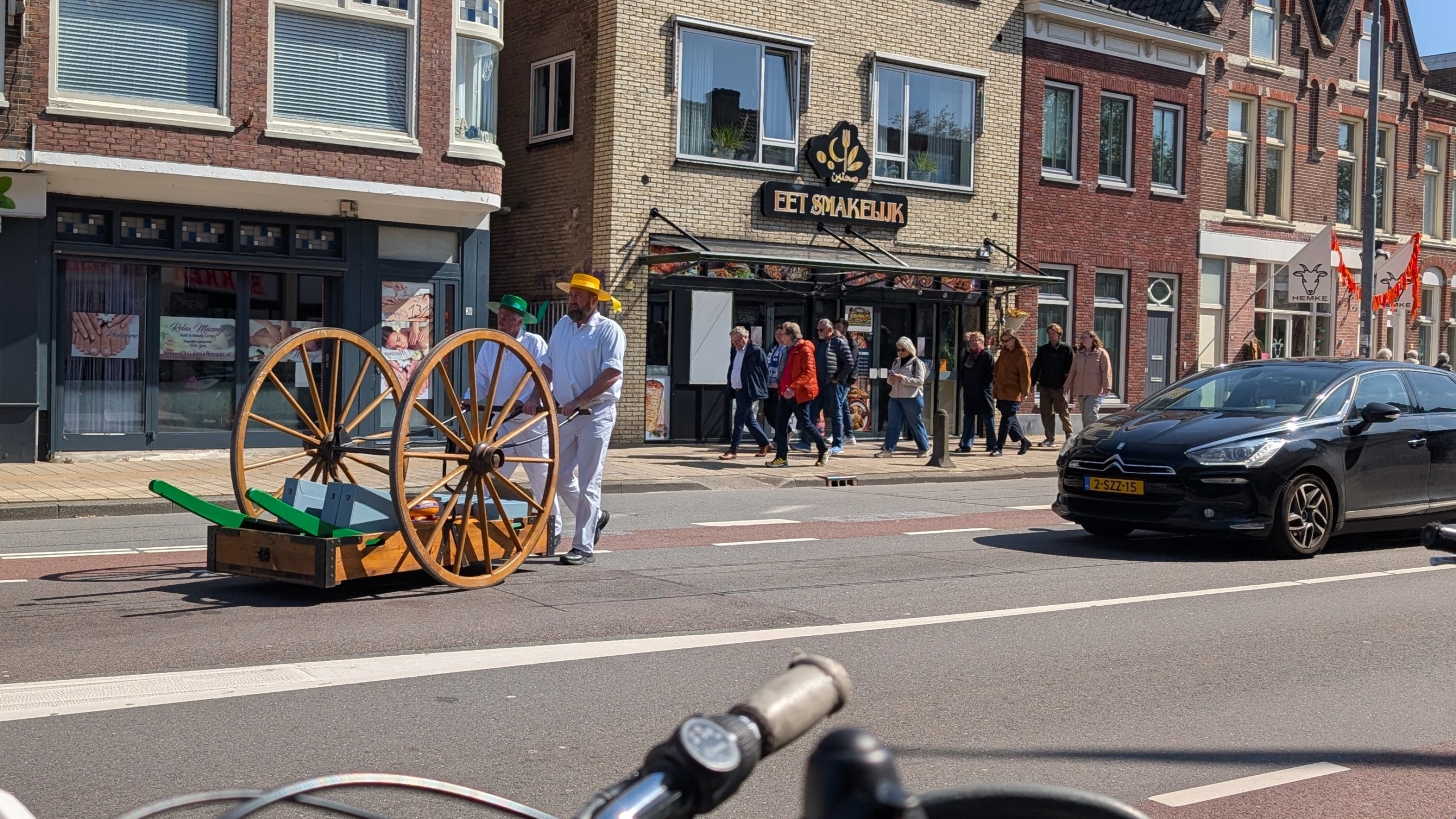 Twee mannen in witte kleding met gekleurde hoeden duwen een houten kaaskar voor een winkel, omringd door voetgangers en auto's.