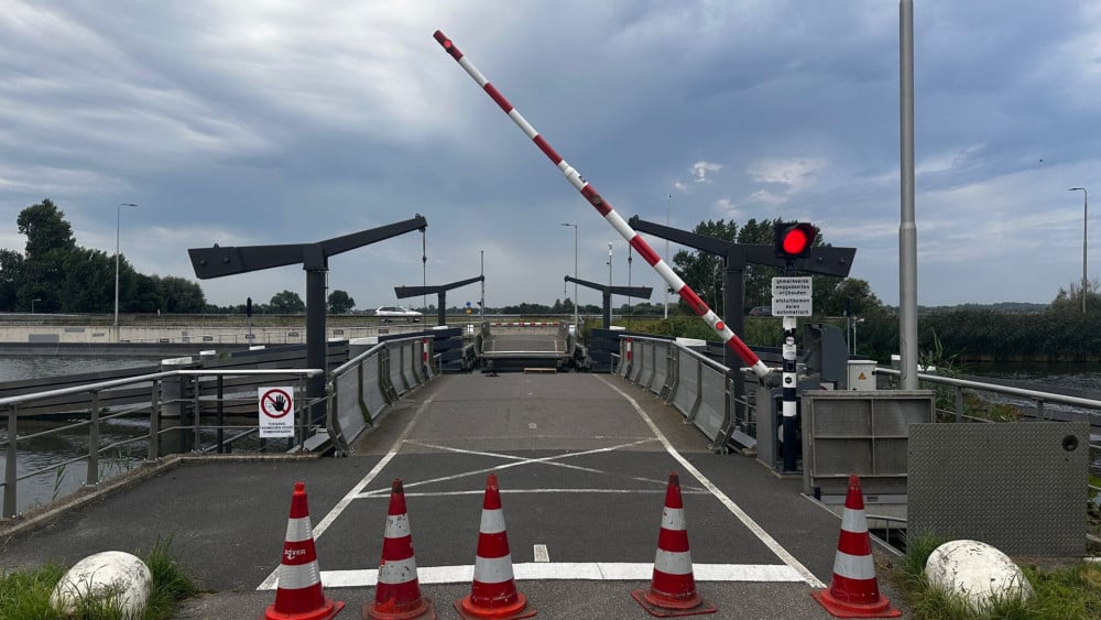Wegversperring bij een open brug met slagbomen omhoog, verkeerslichten op rood en vier verkeerskegels op de voorgrond.