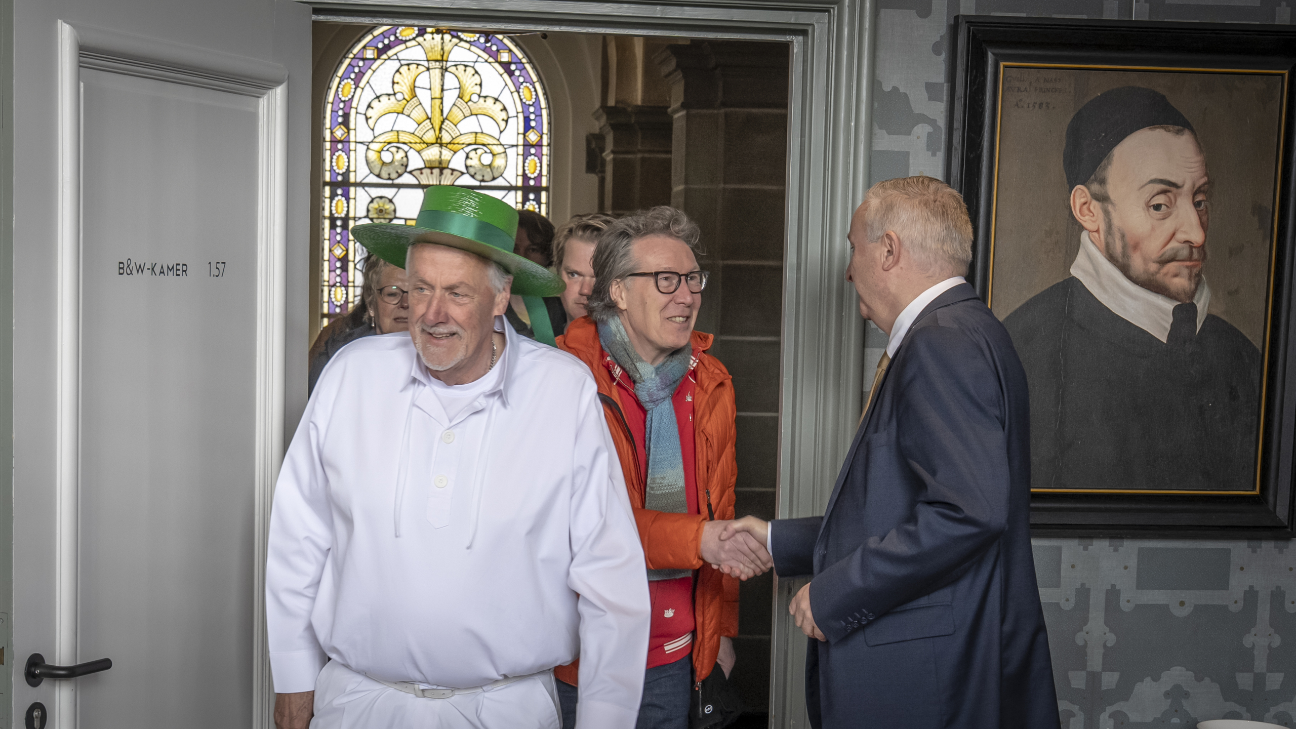 Een man in een witte outfit en groene hoed komt binnen, gevolgd door anderen, terwijl hij een handdruk geeft aan iemand in een pak. Er is glas-in-lood te zien en een schilderij van een historische figuur hangt aan de muur.