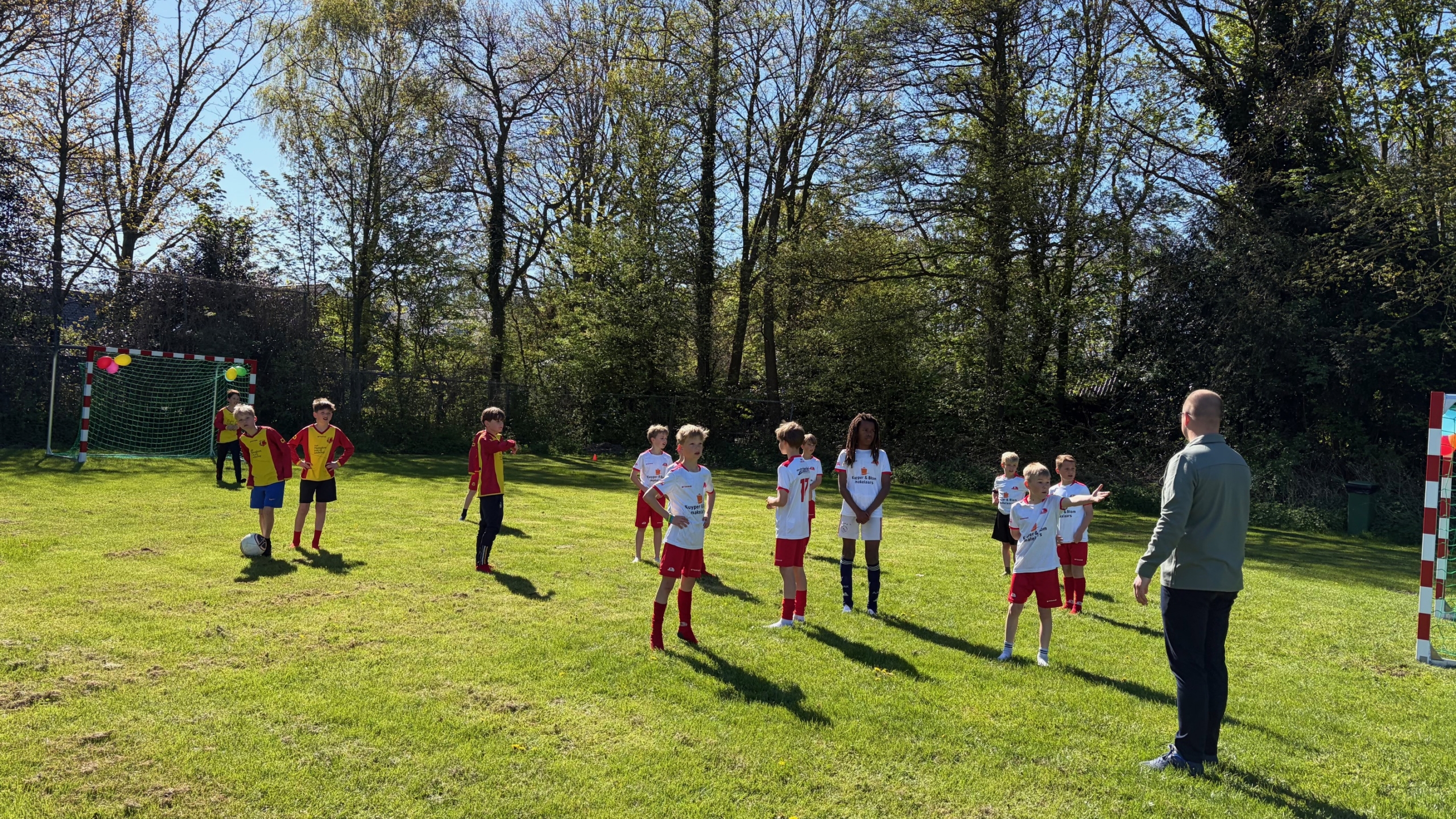 Kinderen spelen voetbal op een grasveld, begeleid door een volwassene bij zonnig weer.