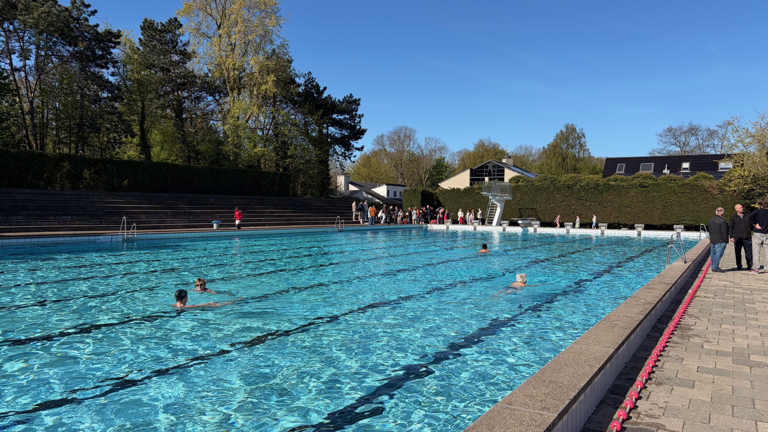 Een buitenzwembad met zwemmers, omgeven door groene bomen en mensen langs de rand, onder een heldere blauwe hemel.