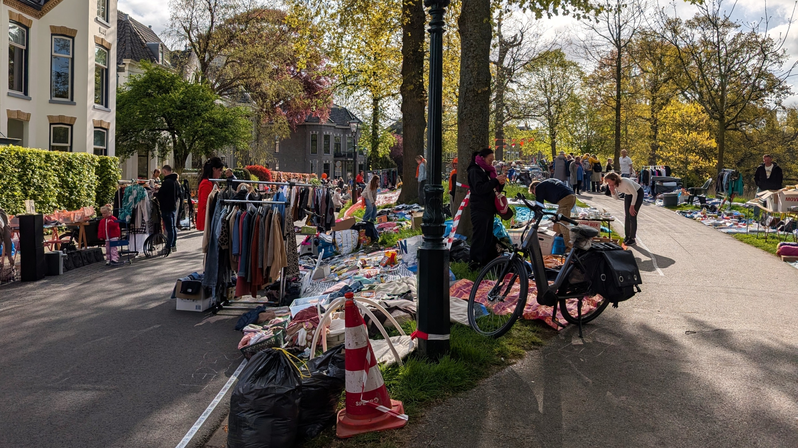 Straatscène van een vrijmarkt met kramen vol kleding en spullen, omgeven door mensen, bomen en huizen.