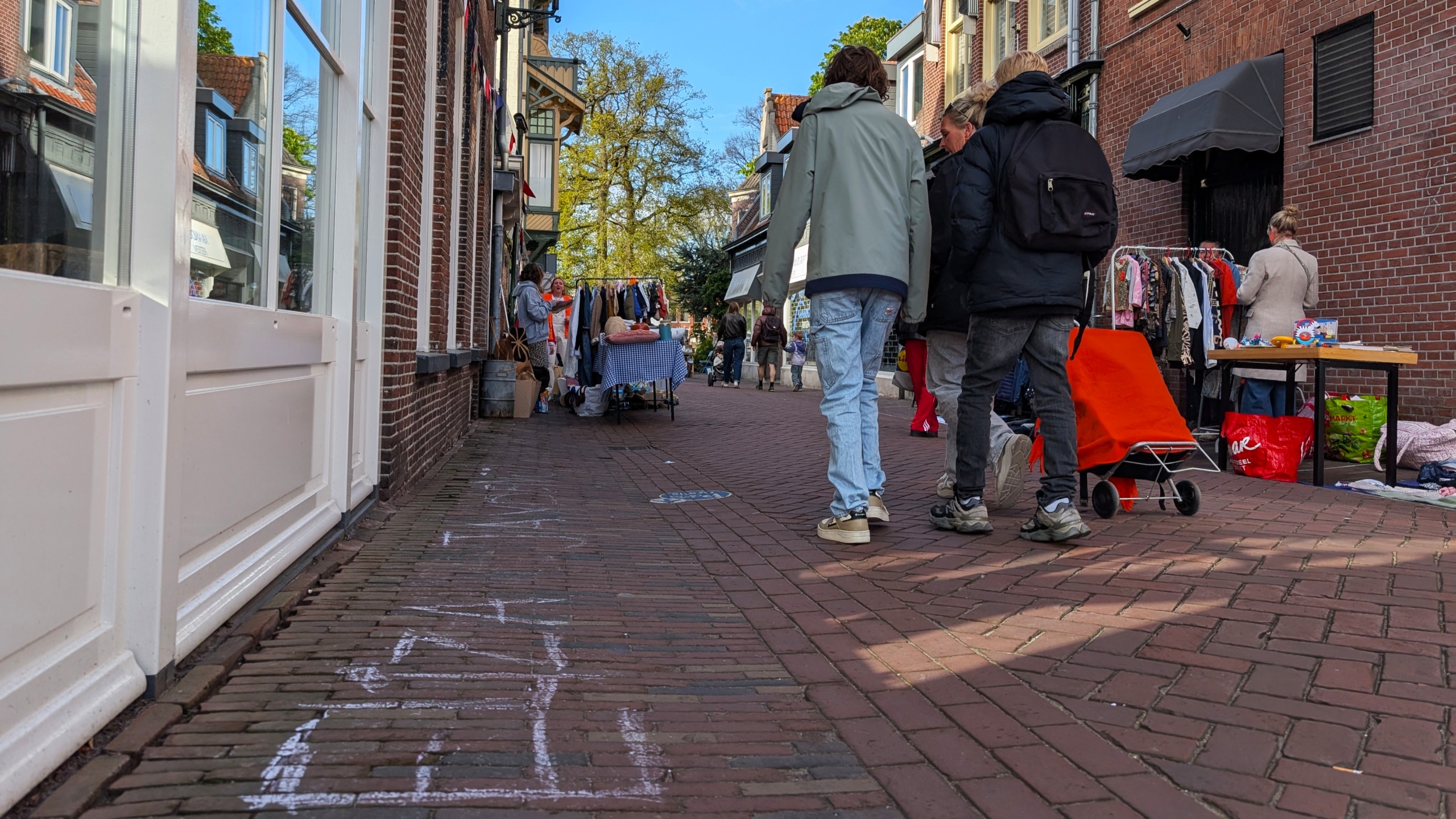Een levendige straat met mensen langs kraampjes met kleding, tafels met verkoopwaar en een zonnige dag.