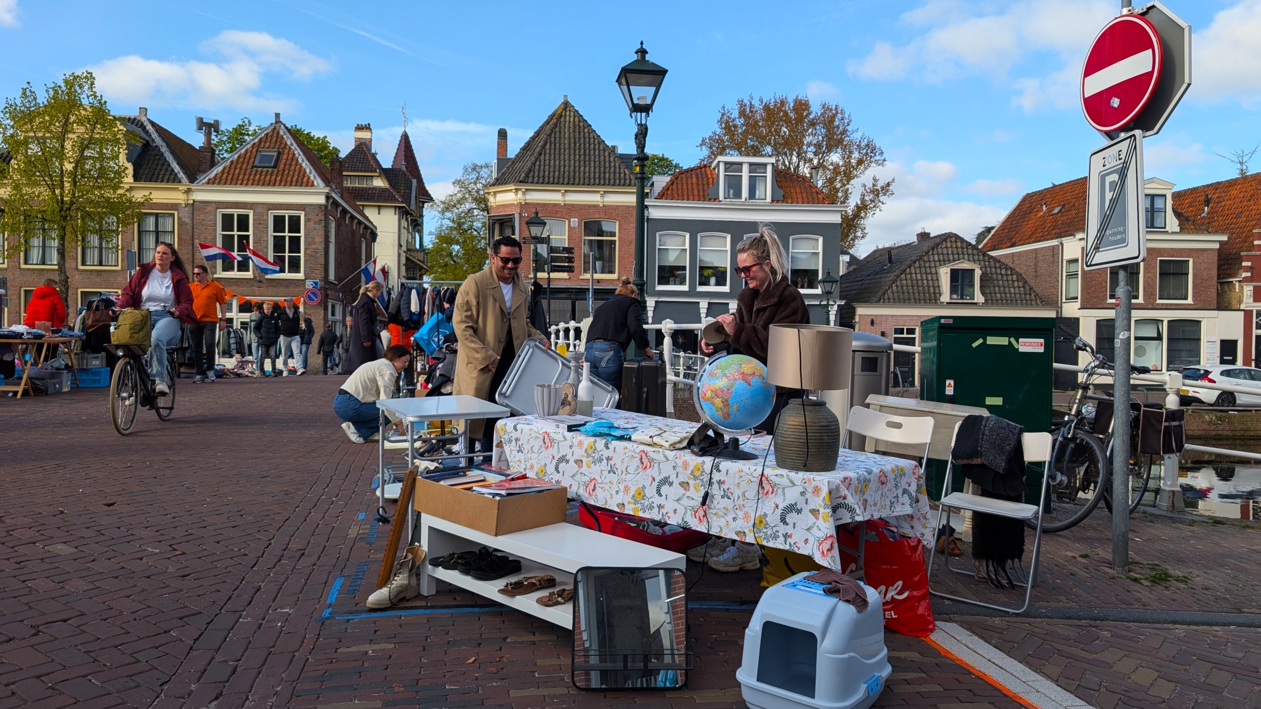 Straatmarkt in een Nederlandse stad met tafels vol tweedehands spullen, mensen die rondlopen en vlaggen aan de achtergrond.