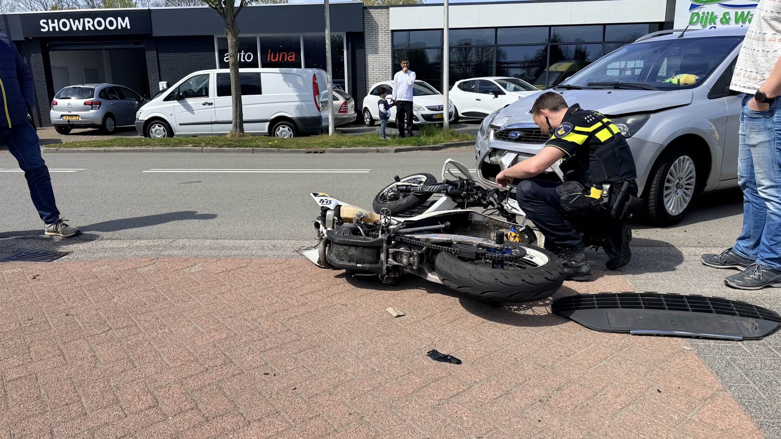 Politieagent onderzoekt een ongeval tussen een motorfiets en een grijze auto, nabij een showroom.