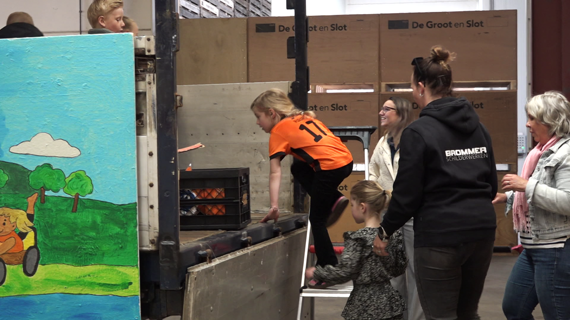 Kinderen en volwassenen bij een vrachtwagen met kleurrijke muurschildering; een kind klimt de laadbak in met hulp.