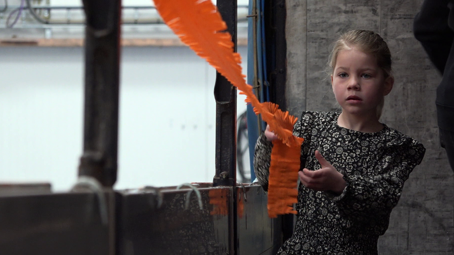 Een jong meisje in een zwart-wit bloemenjurk houdt een oranje papieren slinger vast in een industriële omgeving.