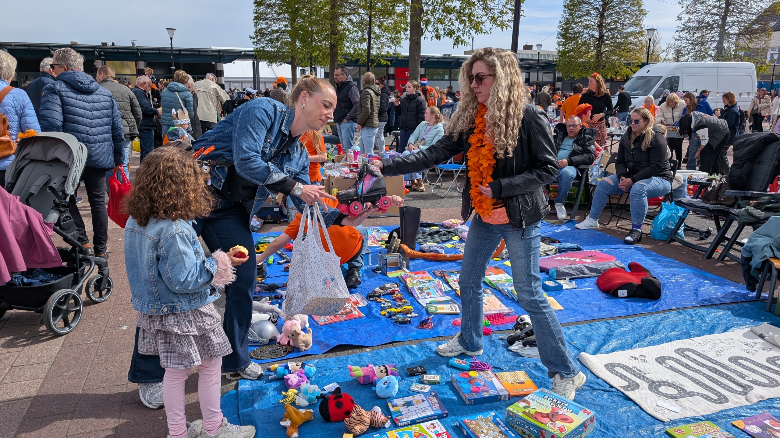 Twee vrouwen ruilen speelgoed op een vrijmarkt terwijl een kind met een appel toekijkt.