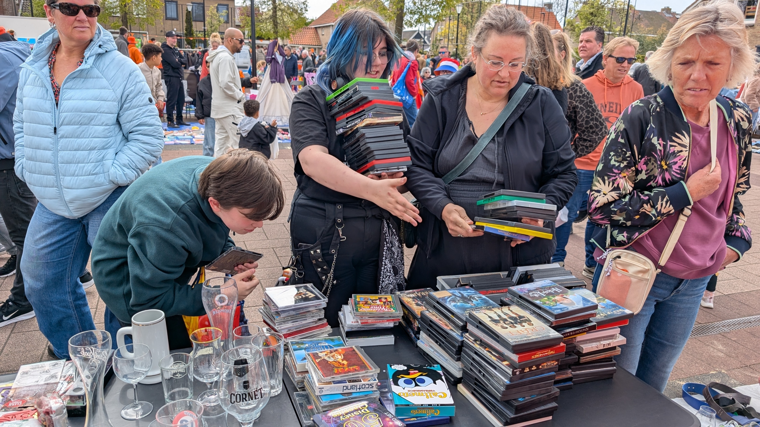 Mensen browsen een markt met tweedehands dvd's op een tafel, omringd door anderen die winkelen.