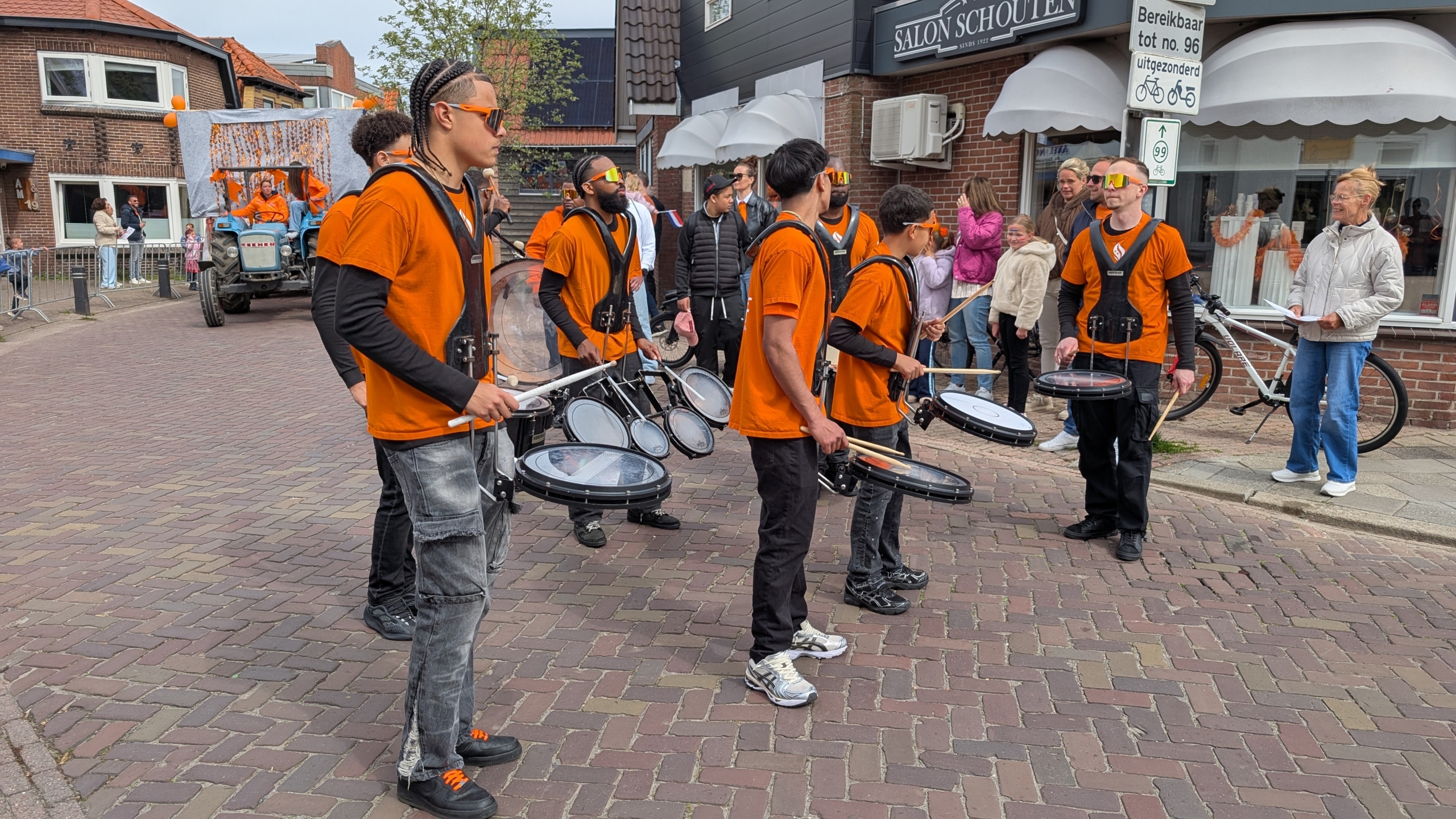Jonge drummers in oranje shirts spelen op straat tijdens een parade, met een versierde tractor op de achtergrond.