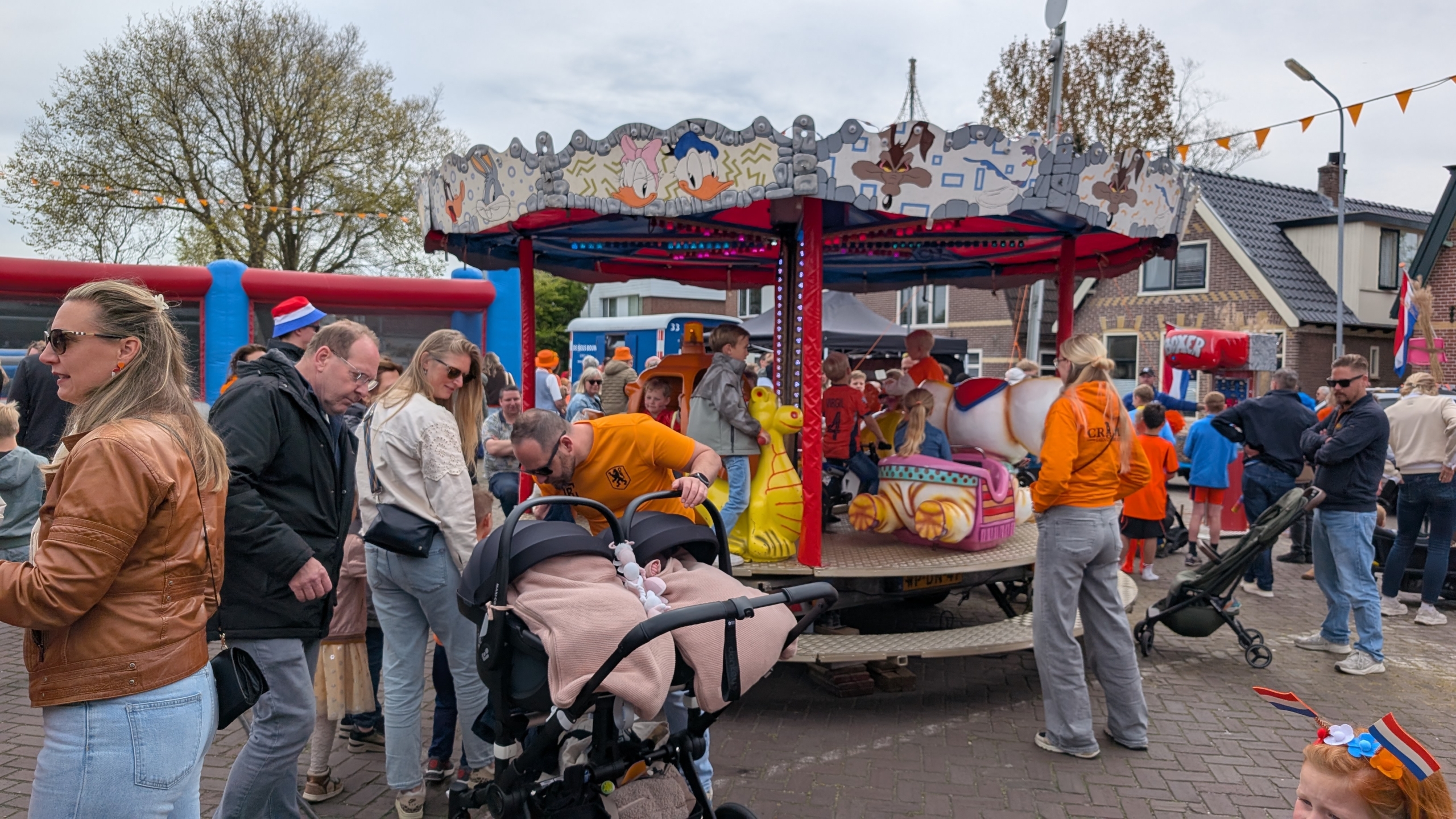 Mensen genieten van een draaiend kinderattractie op een kermis, omringd door feestgangers in oranje kleding.