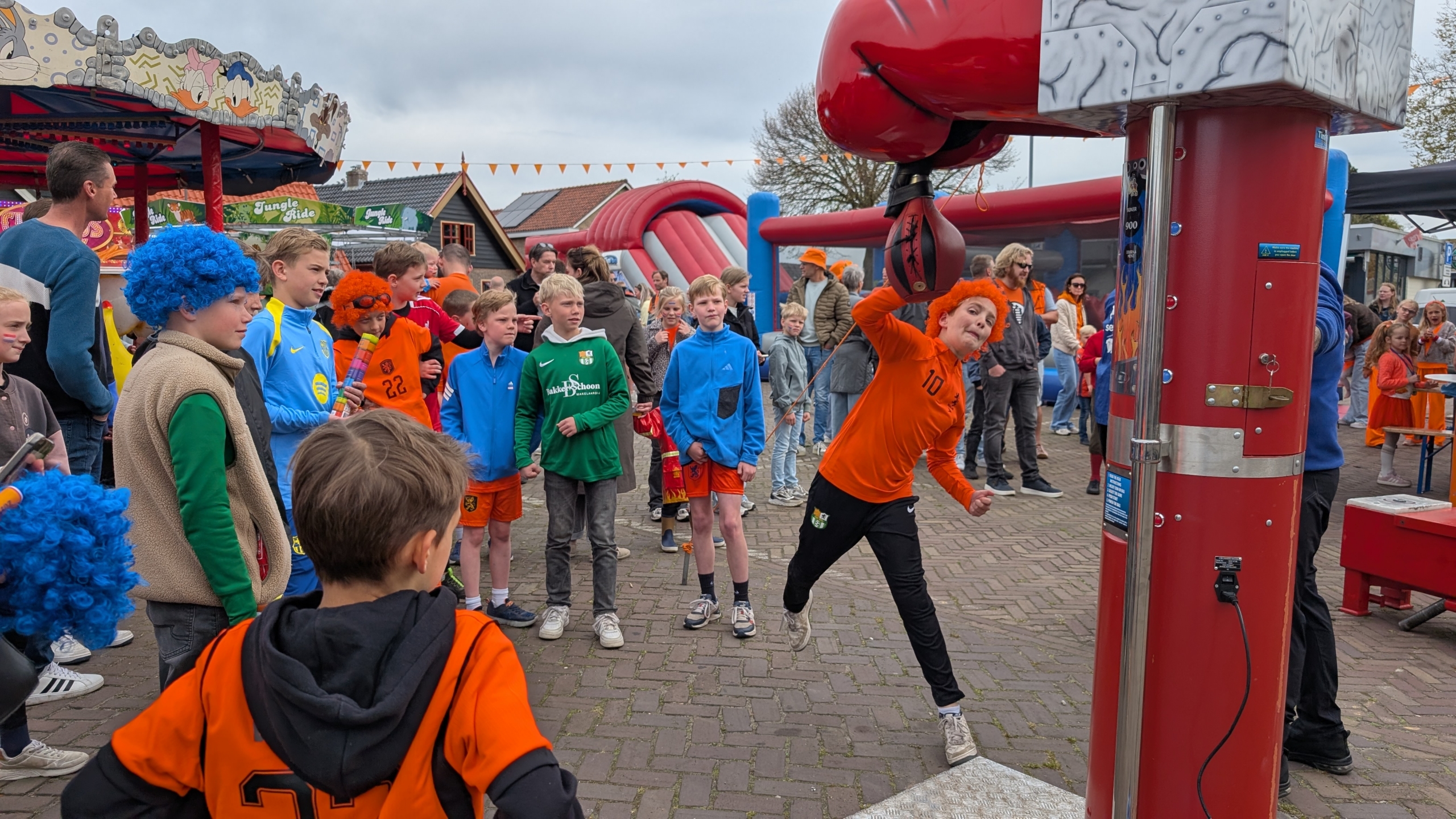 Kinderen in oranje sportkleding kijken naar een jongen die op een bokszak slaat op een kermis.