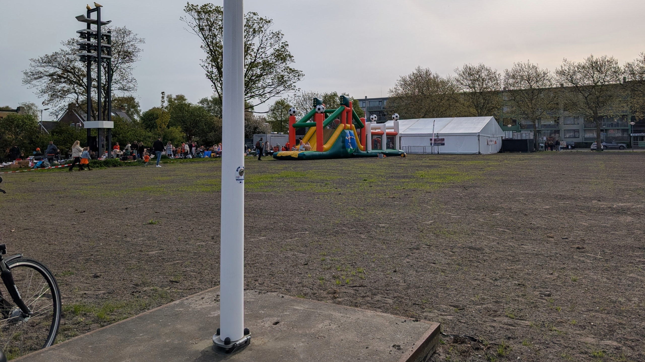 Open terrein met een springkussen en een groep mensen bij een tent, op de achtergrond bomen en woningen.