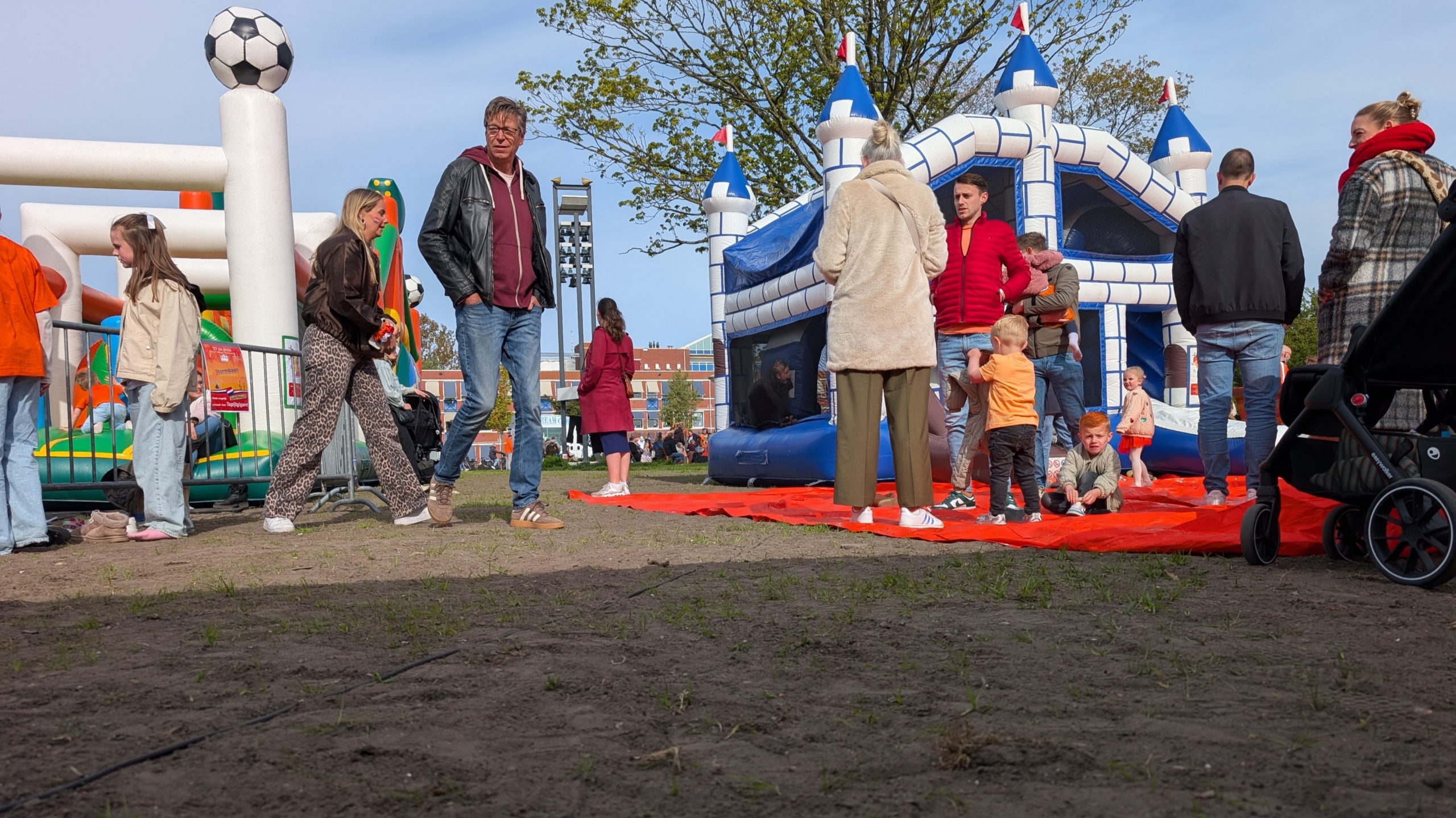 Mensen staan bij springkussens in de vorm van kasteel en voetbal op een buitenevenement, met een kinderwagen op de voorgrond.