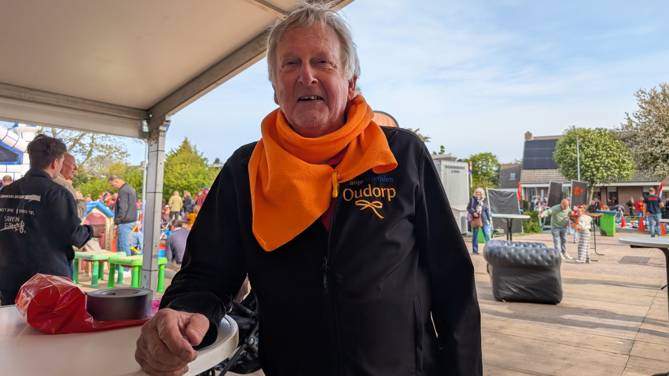 Een oudere man met een oranje sjaal staat onder een tent op een druk feest, met mensen en tafels op de achtergrond.