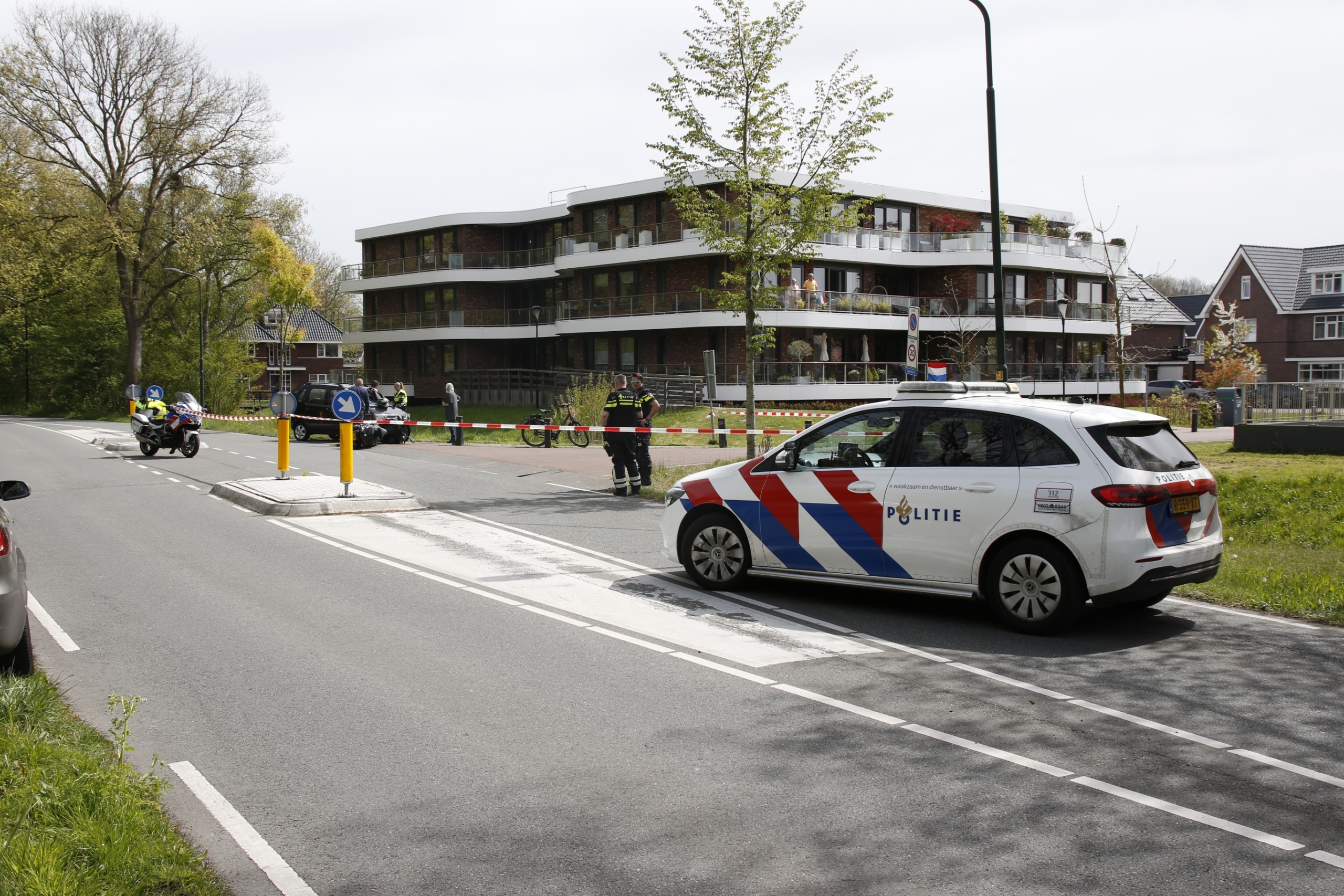 Politievoertuig en motorkap bij afzetting op straat; politieagenten staan bij een groot gebouw.