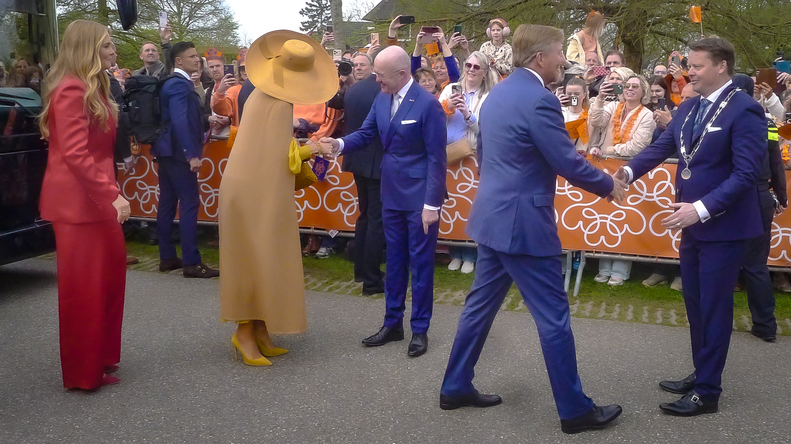 Mensen in chique kleding schudden handen op een openbare bijeenkomst met veel omstanders en oranje decoratie op de achtergrond.