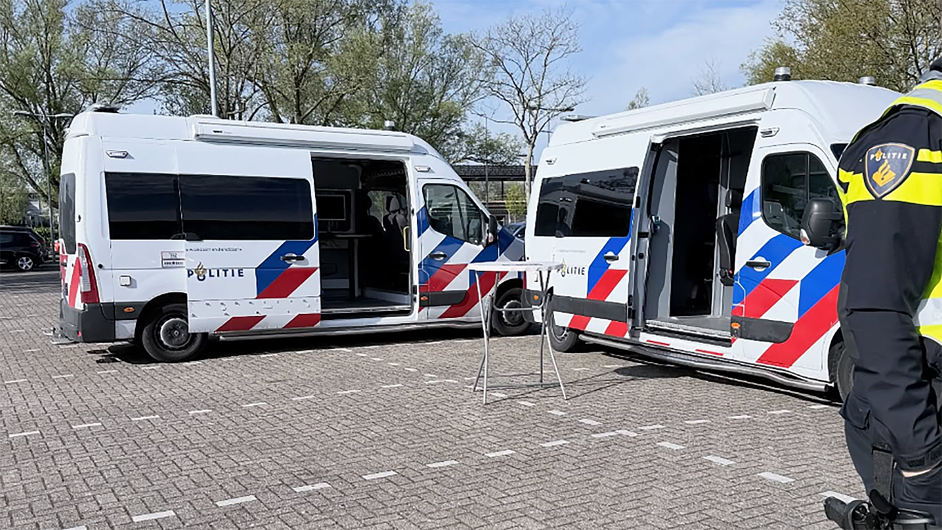 Twee geopende politiebusjes staan geparkeerd op een plein, met een agent zichtbaar aan de rechterkant.