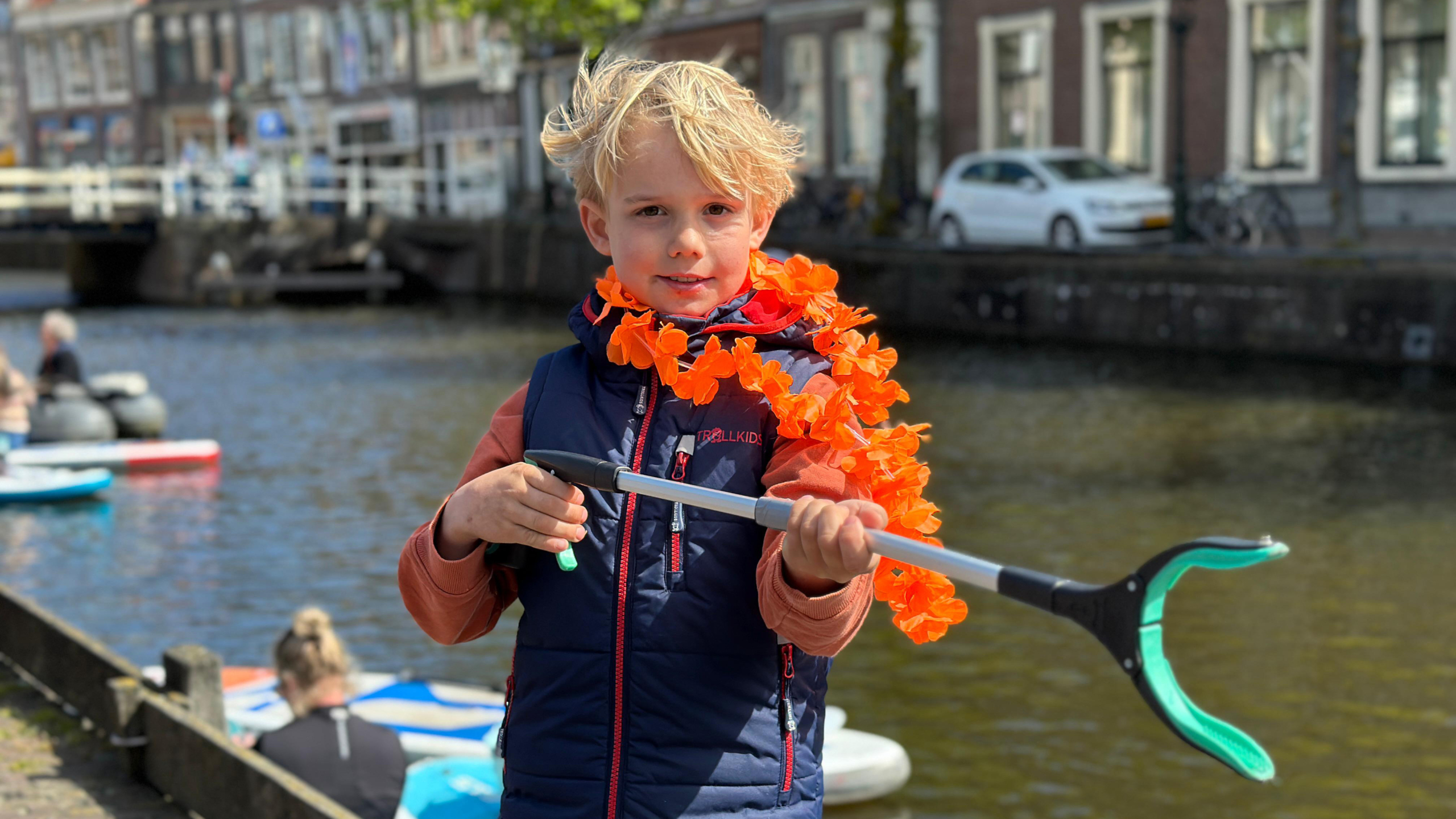 Jonge jongen met oranje bloemenkrans houdt grijper vast bij een kanaal, met gebouwen en water op de achtergrond.