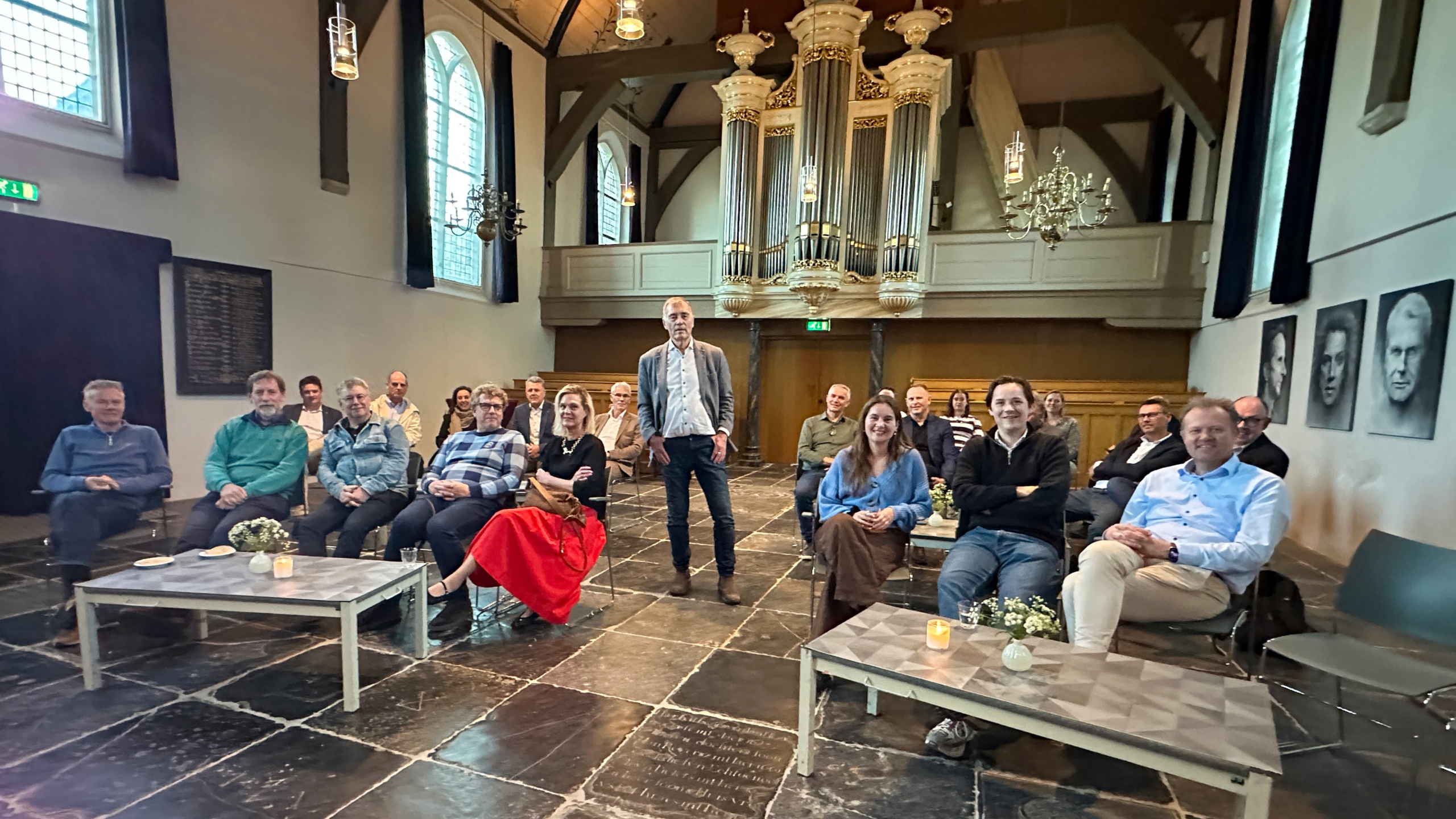 Groep mensen zittend in een kerkachtige ruimte met een groot orgel op de achtergrond, ontspannen en glimlachend.