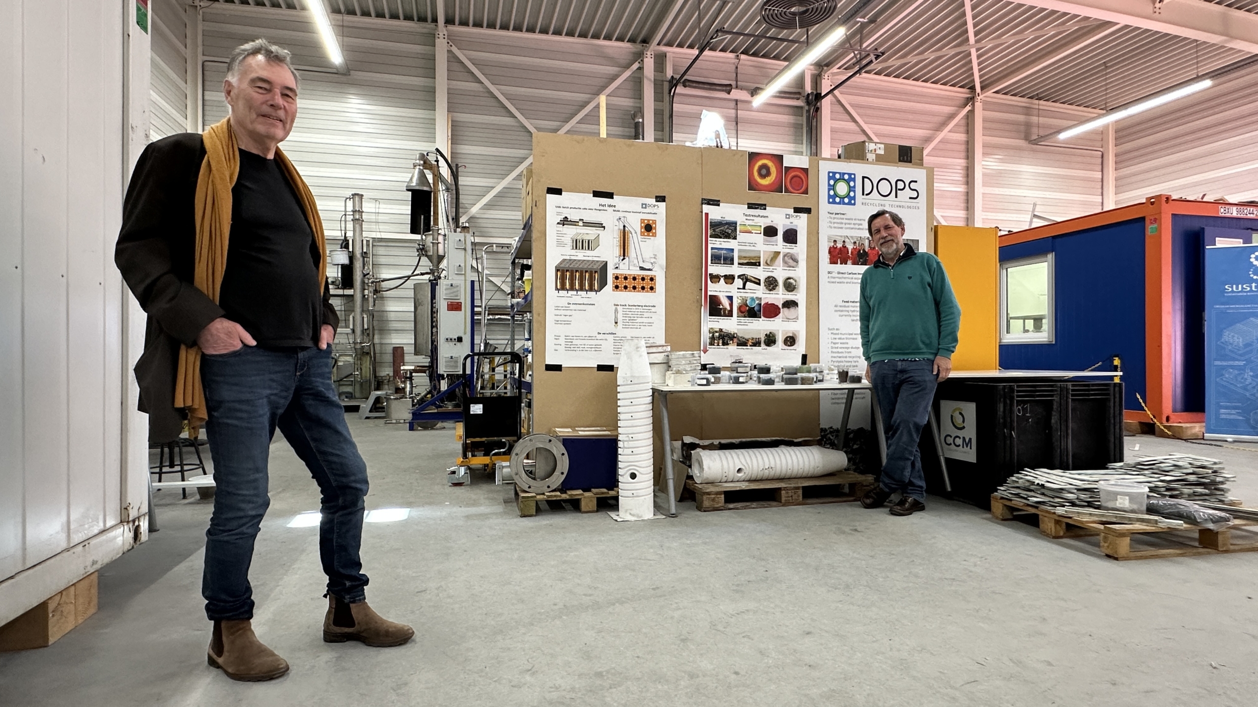 Twee mannen staan in een werkplaats bij een tentoonstellingspaneel over recyclingtechnologie, omringd door apparatuur.