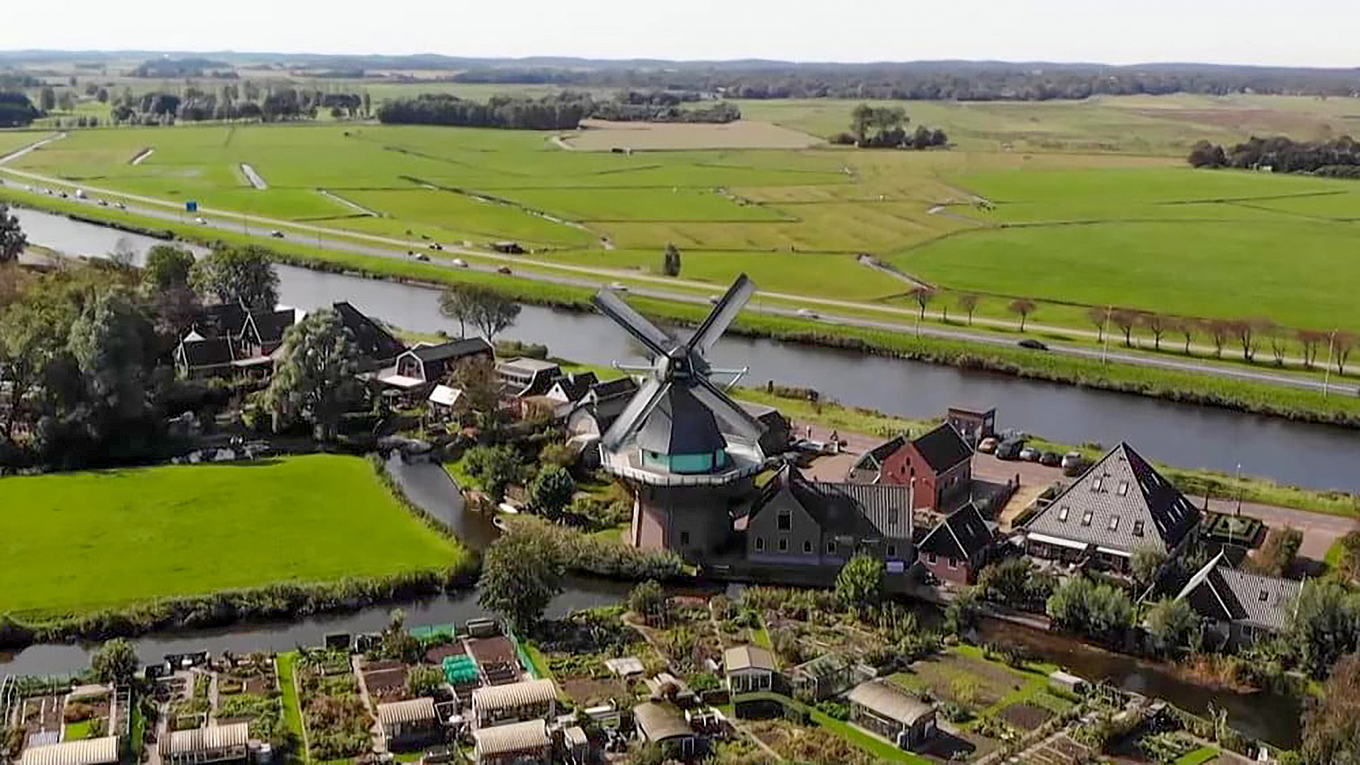 Luchtfoto van een dorp met een traditionele windmolen, omringd door groene velden en een kanaal.