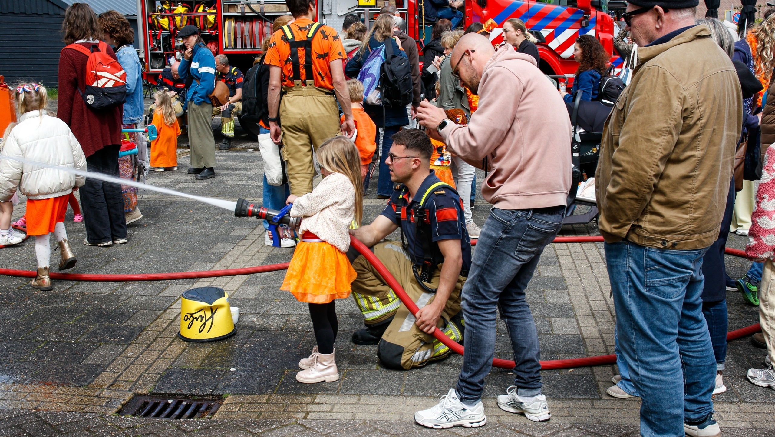 Een kind in een oranje rok spuit water met een brandslang onder begeleiding van een brandweerman tijdens een evenement.