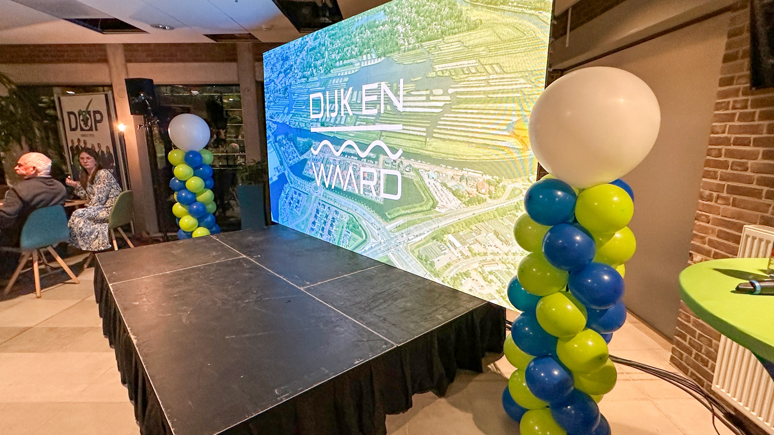 Podium met ballonnen in blauw en groen bij een groot scherm met de tekst "DIJK EN WAARD", naast een tafel met mensen aan de zijkant.