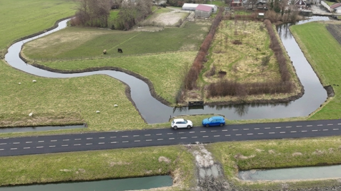 Twee auto's op een weg naast een bochtige sloot en weiland met koeien, gefotografeerd vanuit de lucht.